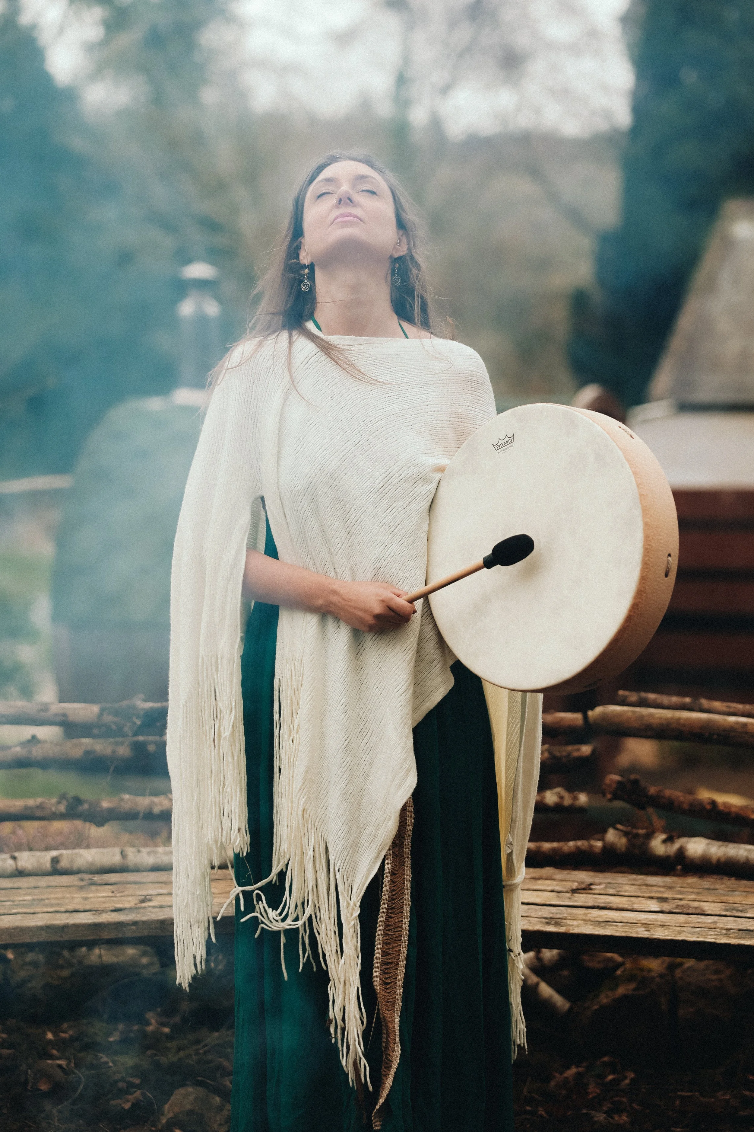 A woman dressed in traditional Native American attire standing outdoors with her eyes closed, holding a large, round drum and a drumstick, surrounded by logs and trees.