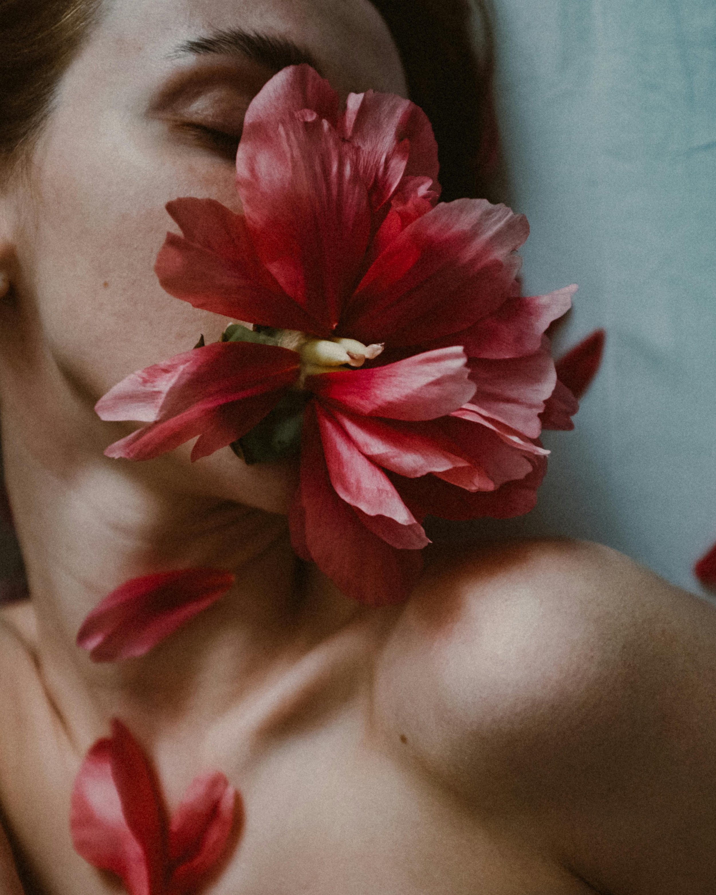 A close-up of a person's face with closed eyes, holding a vibrant red flower close to their nose, showing detailed petals and a hint of their shoulder.