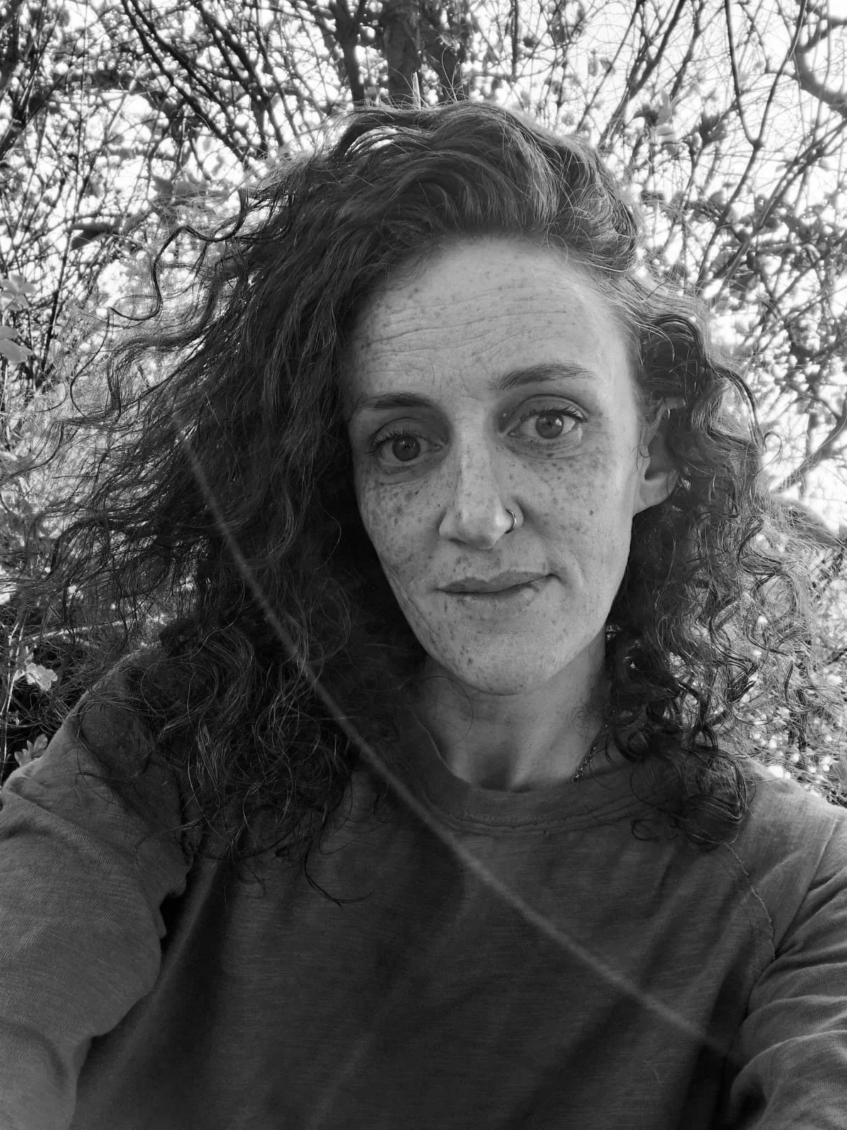 Black and white photo of a woman with curly hair and freckles, taking a selfie outdoors with trees in the background.