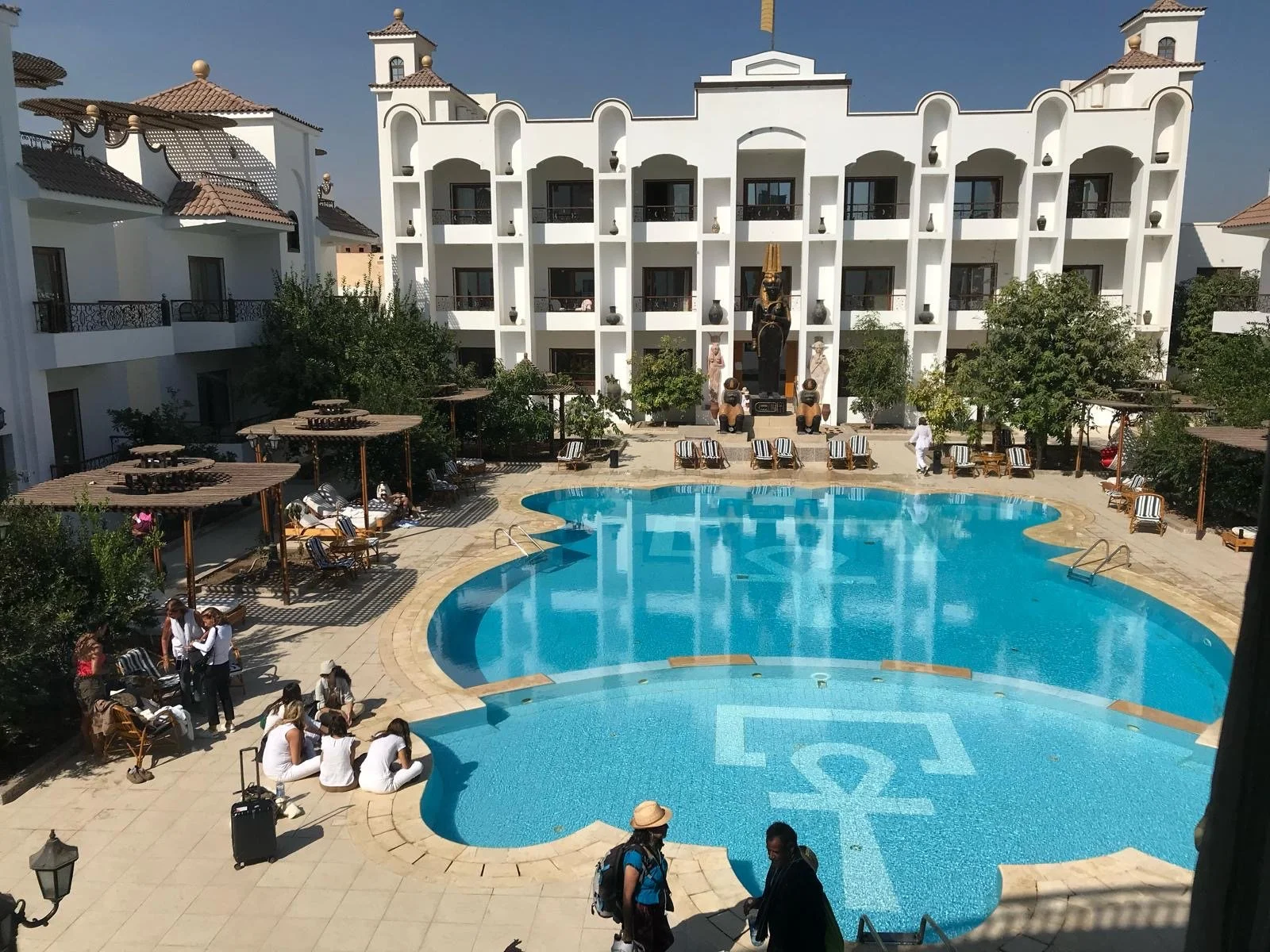 A luxury hotel courtyard with a swimming pool, surrounded by lounge chairs, umbrellas, and lush greenery, with a white multi-story building in the background.