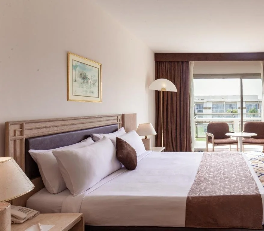 Hotel room with a large bed, beige headboard, white sheets, and brown accent pillows. There are bedside lamps, a painting above the bed, a seating area with two chairs, a table, and a sliding glass door with curtains leading to a balcony with a view of a building and greenery.