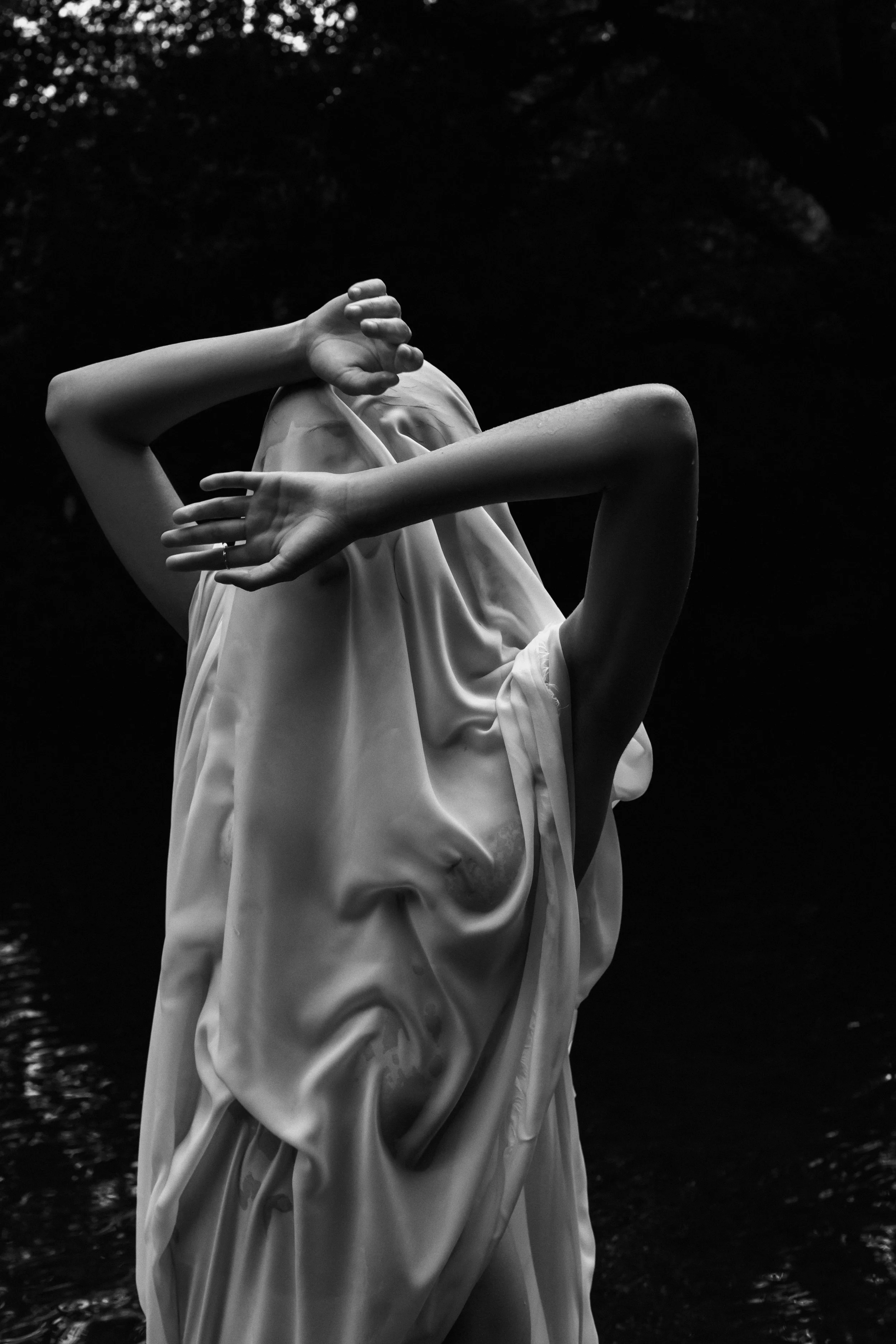 Black and white photograph of a sculpture of a woman with her hand covering part of her face, standing near water with a dark, blurred background.