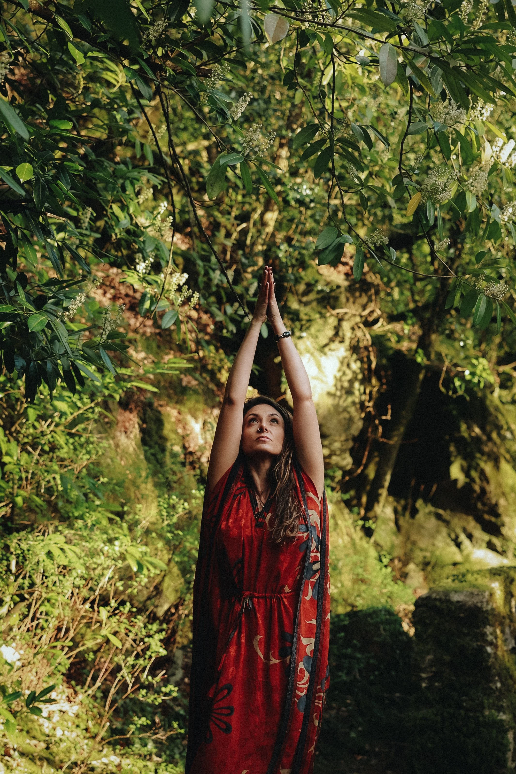 A woman standing outdoors among green trees with her arms raised above her head, hands pressed together, wearing a red and black dress with floral patterns, in a natural forest setting.
