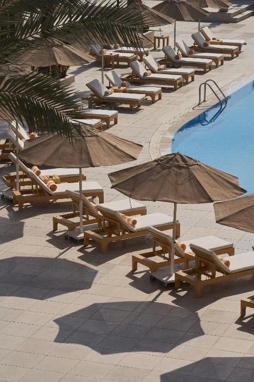 Empty poolside lounge chairs with beige umbrellas and rolled towels next to a swimming pool at a resort or hotel.