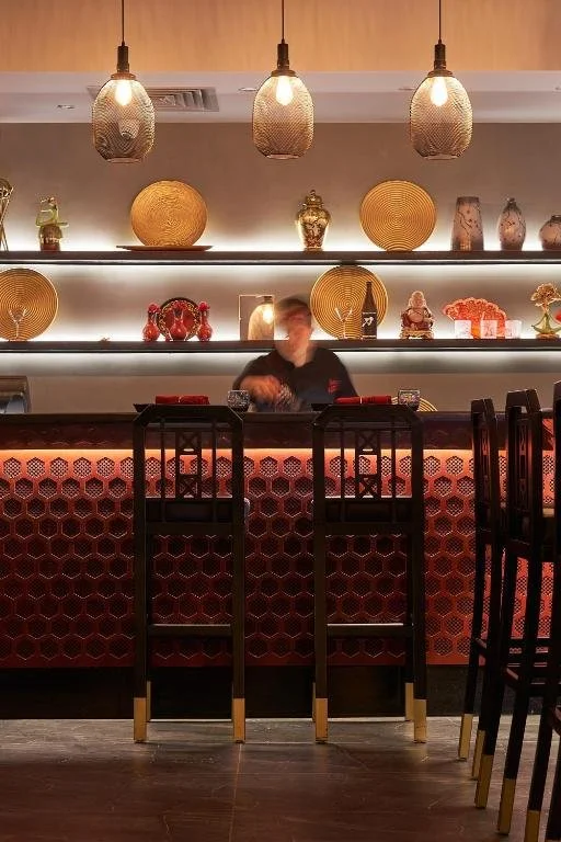 Interior of a modern bar or restaurant with a decorative counter, hanging pendant lights, and shelves with vases and decorative objects in the background.