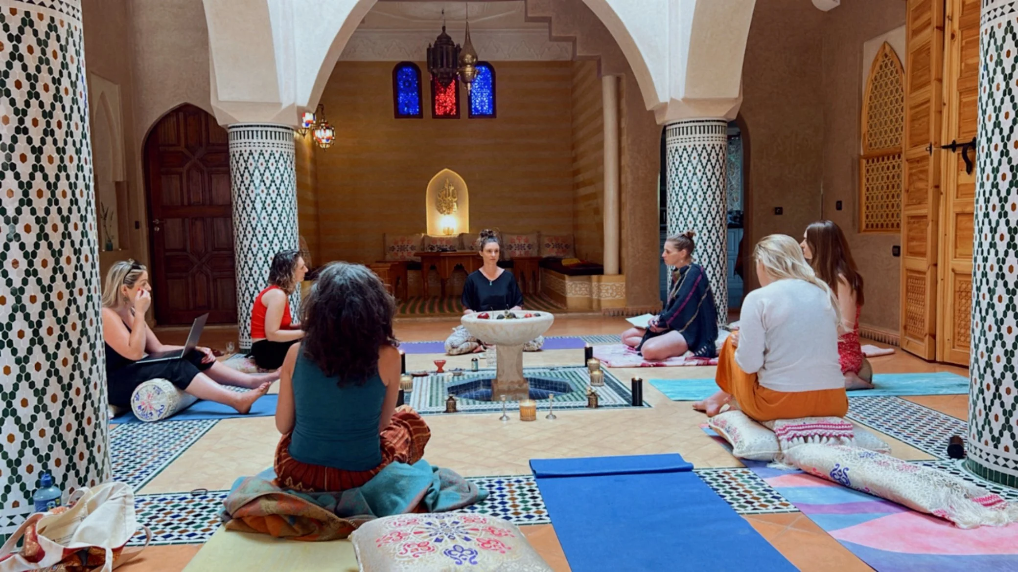 Group of women participating in a meditation session in a room with Moroccan-style decor, including intricate tile patterns, draped fabrics, and a central fountain.