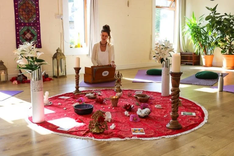 A woman in a white blazer and black top sitting cross-legged on a red circular rug in a cozy, well-lit room. She is engaged with a wooden box placed in front of her with various spiritual objects, including candles, flowers, and statues, arranged on the rug. Yoga mats and green cushions are placed around the room, alongside potted plants and decorative items like lanterns and flowers.