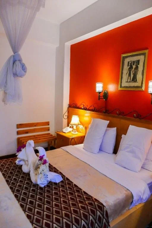 A cozy hotel room with a bed that has three pillows, a blanket, and decorative towel swans on the bed. The room features a red accent wall, a wooden headboard, a nightstand with a lamp, a piece of framed artwork, and wall sconces for lighting. There is a small wooden bench and a sheer curtain tied back on the window.