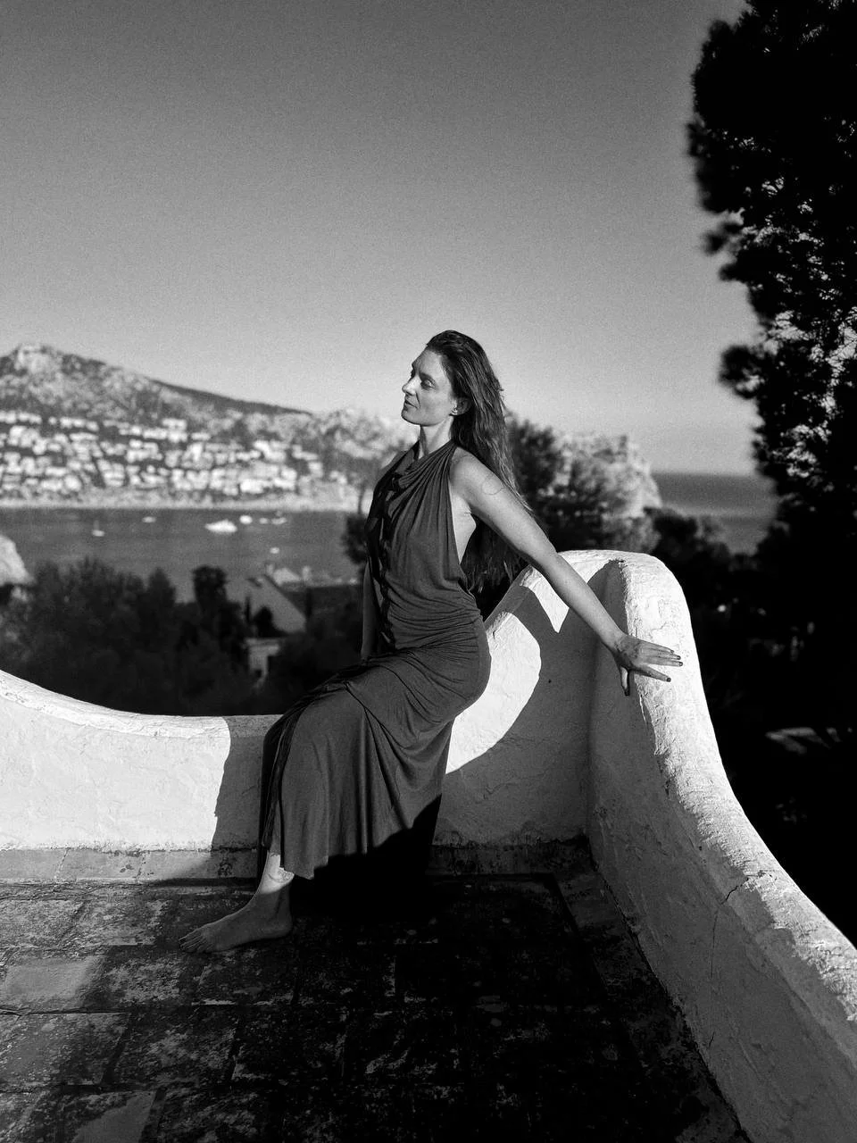 A woman in a long dress is sitting on a curved stone wall outdoors, with a body of water and hillside town in the background.