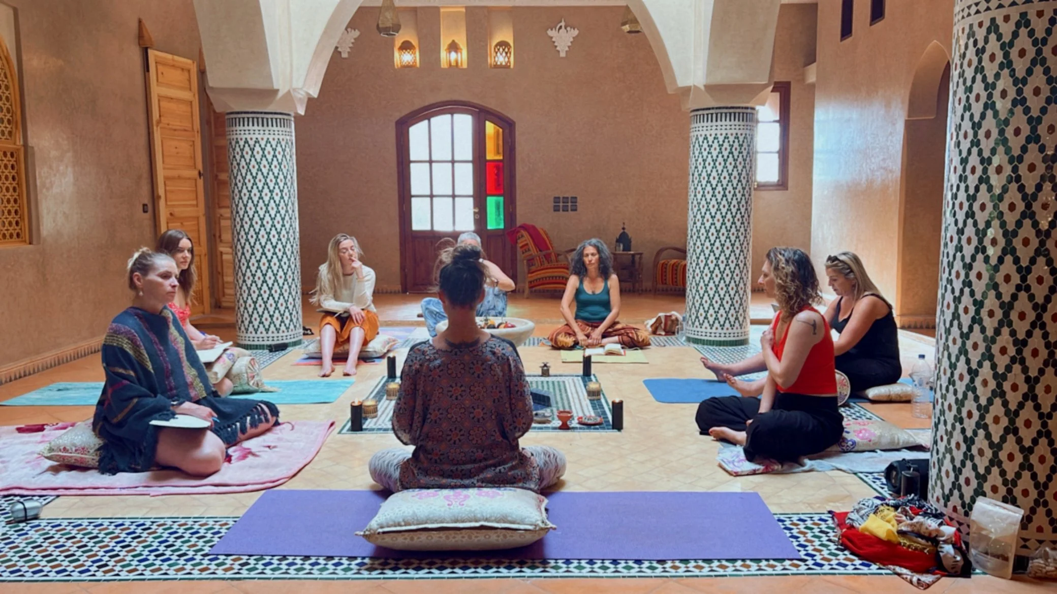 A group of women sitting cross-legged on cushions and yoga mats in a room with Moroccan decor, including mosaic columns and colorful stained-glass windows, participating in a spiritual or meditation session.