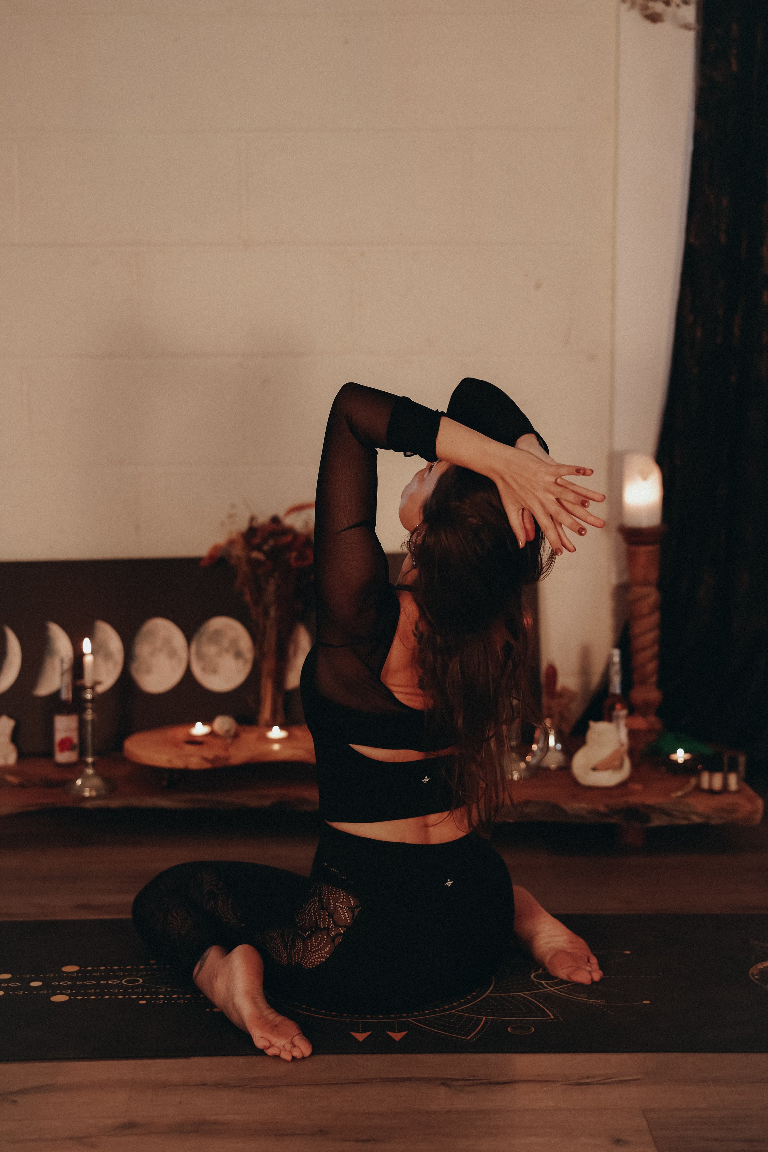 A woman practicing yoga or dance indoors at night, surrounded by candles, flowers, and decorative items, wearing black leggings and a sheer black top.