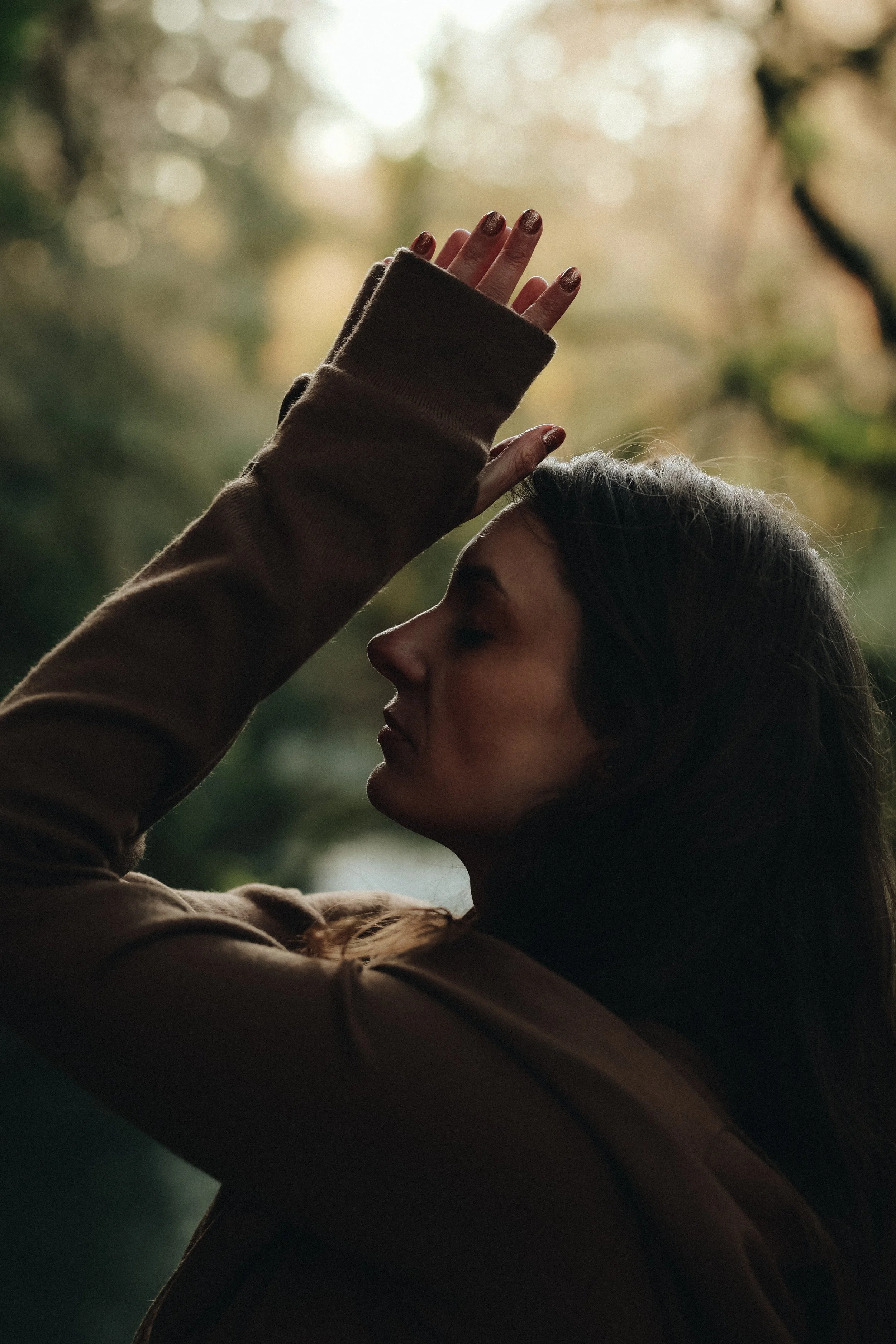 A woman with long dark hair and closed eyes is touching her forehead with her hand, which has painted nails, wearing a brown long-sleeve shirt. The background is blurred outdoors with sunlight filtering through trees.