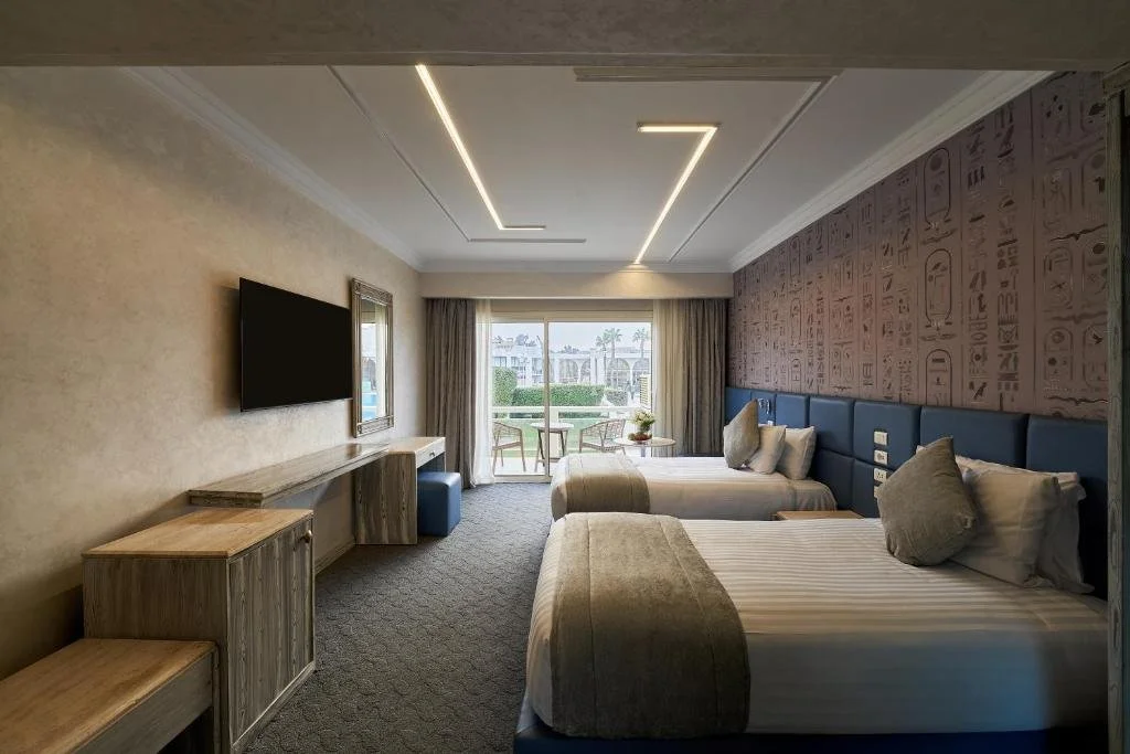Hotel room with two beds, a wall-mounted TV, a balcony with outdoor furniture, and a decorative wallpapered wall.