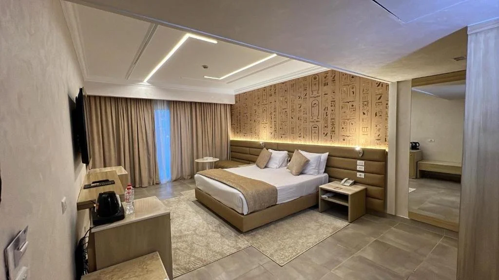Modern hotel room with a large bed, beige headboard, and Egyptian hieroglyphics wall art. There is a TV on the wall, a small desk with a water bottle and kettle, and a window with beige curtains.
