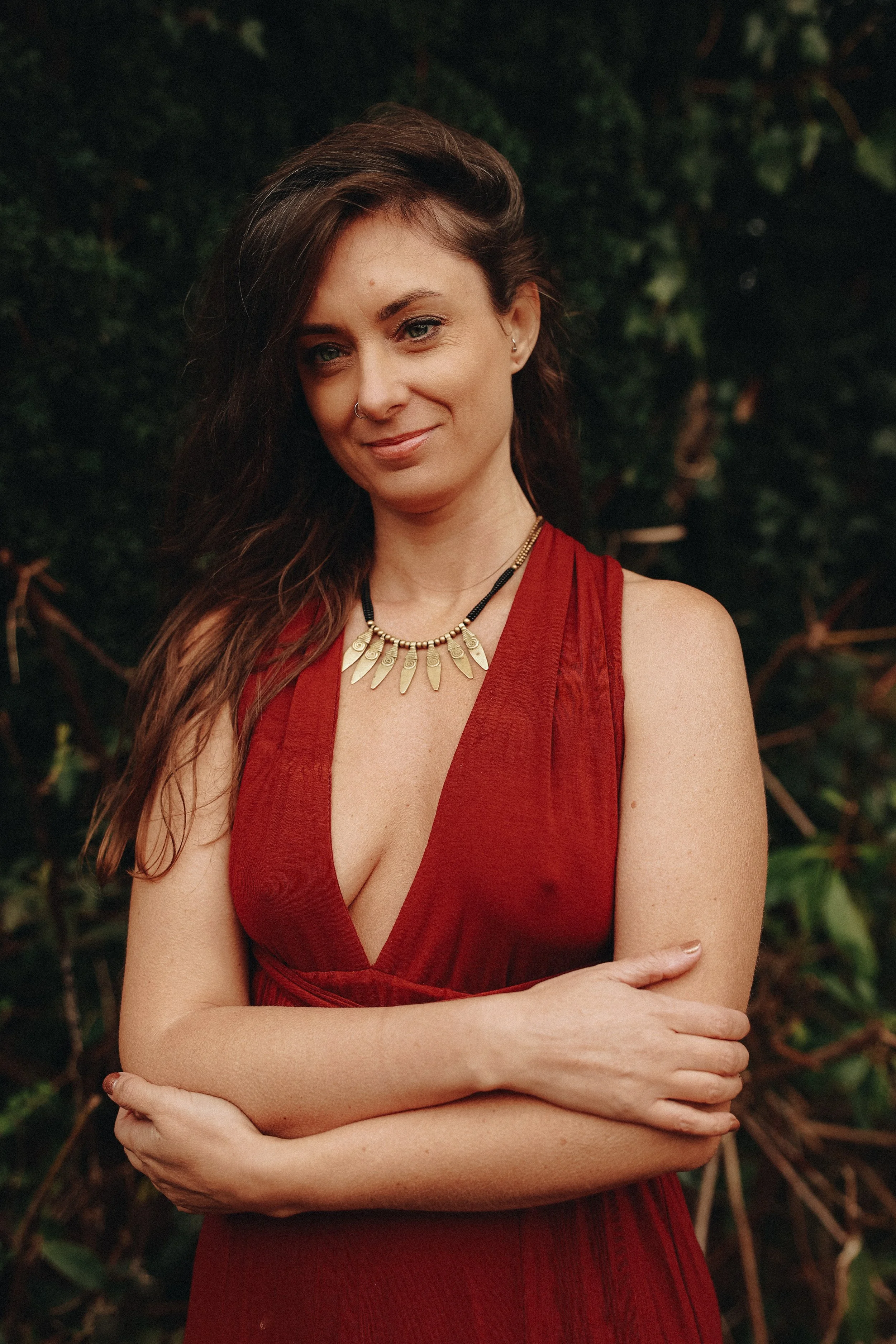 A woman with long, wavy brown hair wearing a red sleeveless dress and a decorative necklace, standing outdoors with green foliage in the background.
