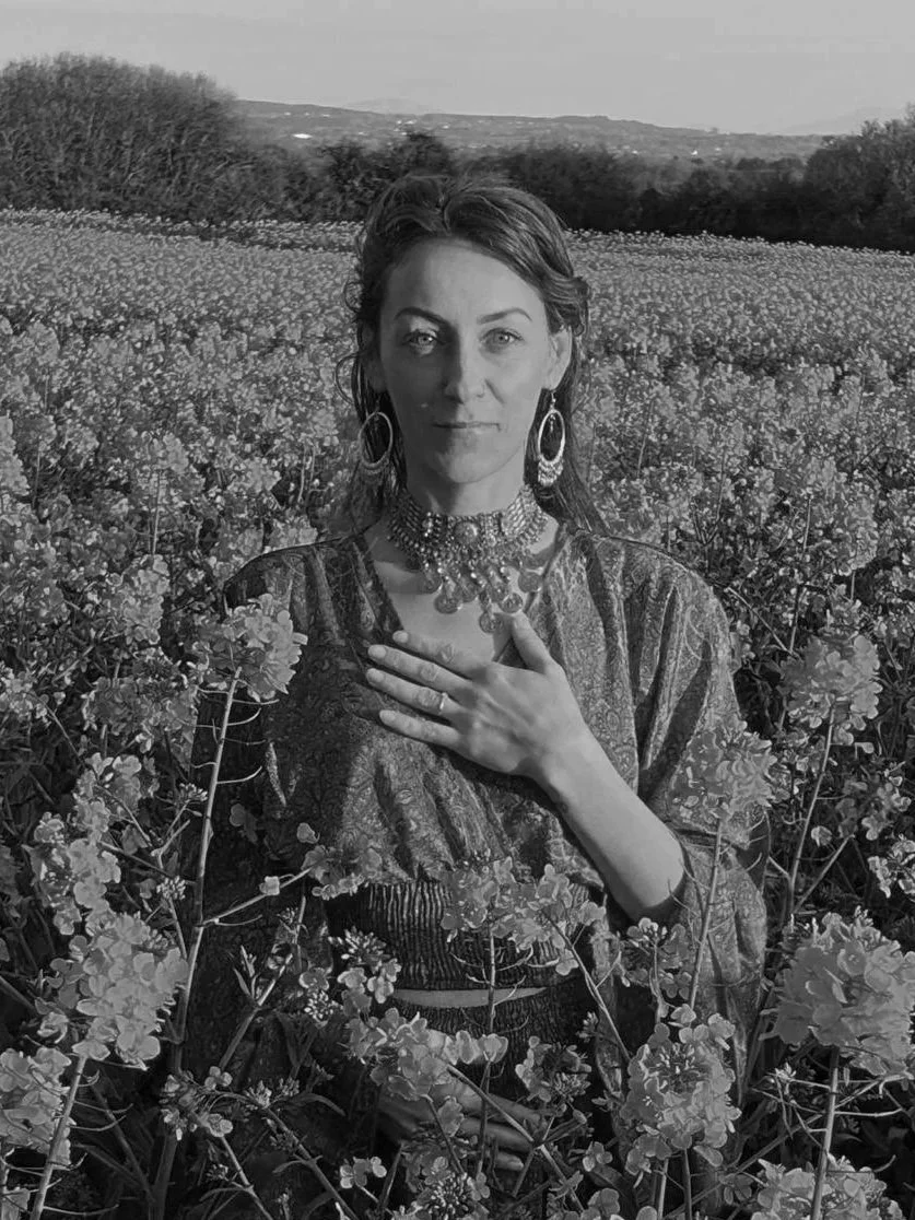 Black and white photo of a woman standing in a field of flowers, wearing jewelry, with her hand over her chest, looking directly at the camera.