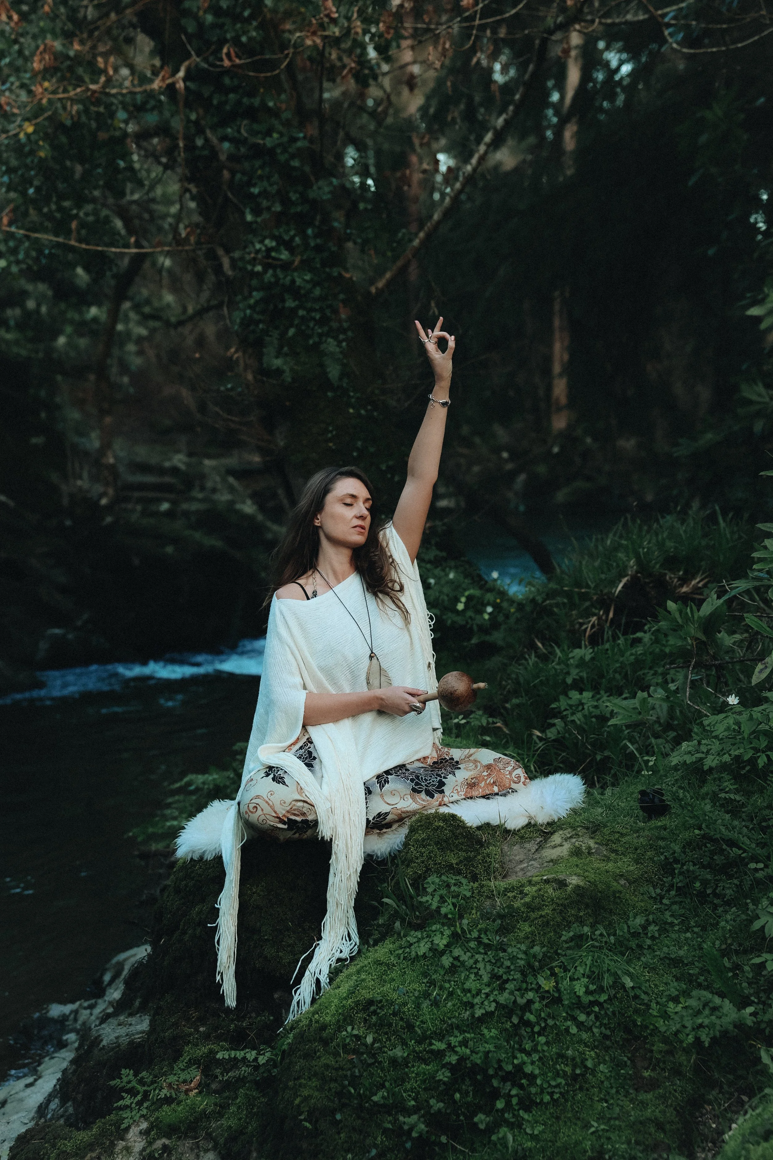 A woman meditating outdoors near a river in a lush green forest. She is sitting cross-legged on a furry white mat, holding a rattle, with her eyes closed and a peaceful expression. She is making a gesture with her right hand, and wearing a loose white top, patterned pants, and various jewelry.