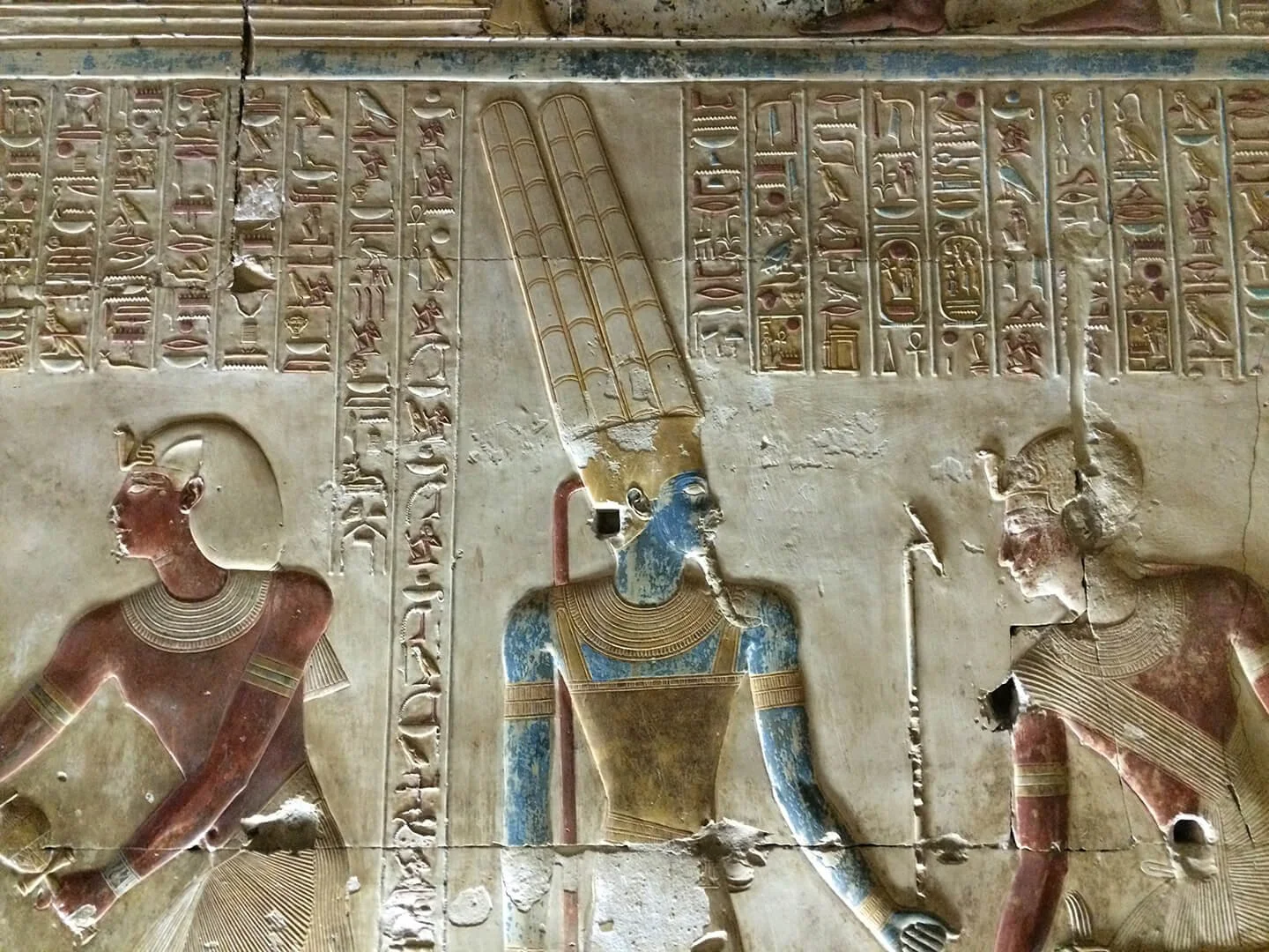 Ancient Egyptian wall carving with hieroglyphs and figures, including a central figure wearing a tall headdress, with two other figures on either side, all depicted in profile.