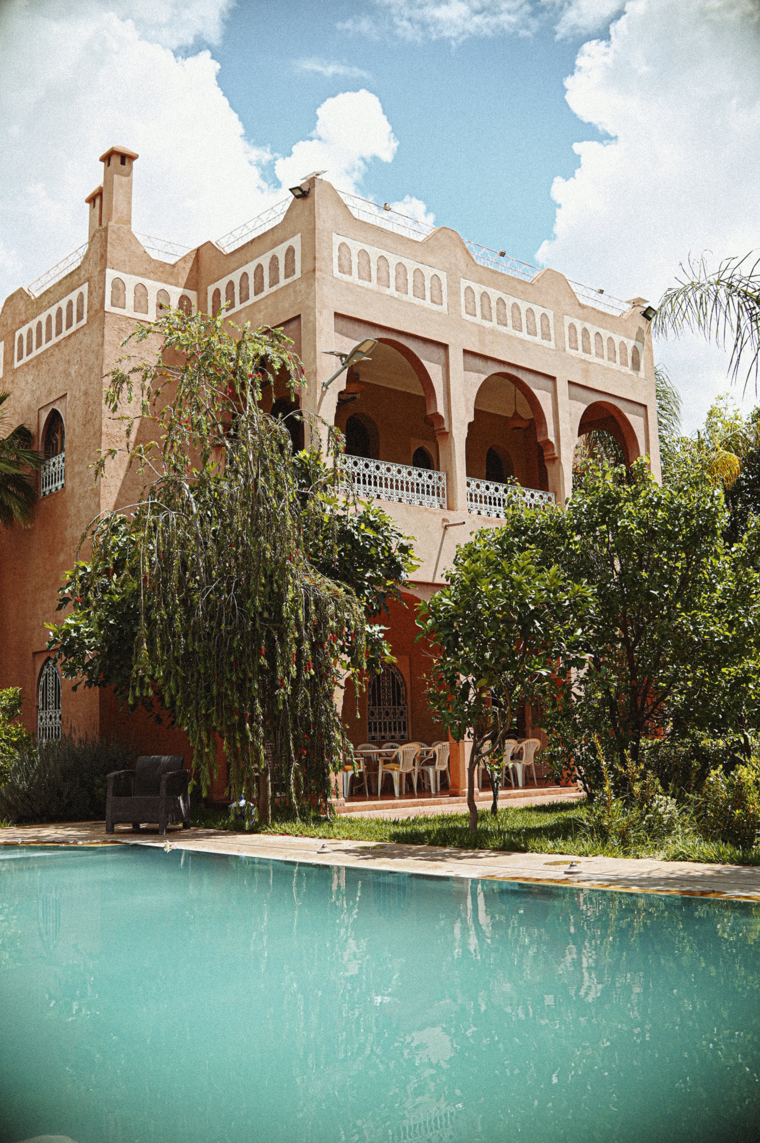 A large house with arched windows and balconies, surrounded by trees, with a swimming pool in the foreground and a partly cloudy sky above.
