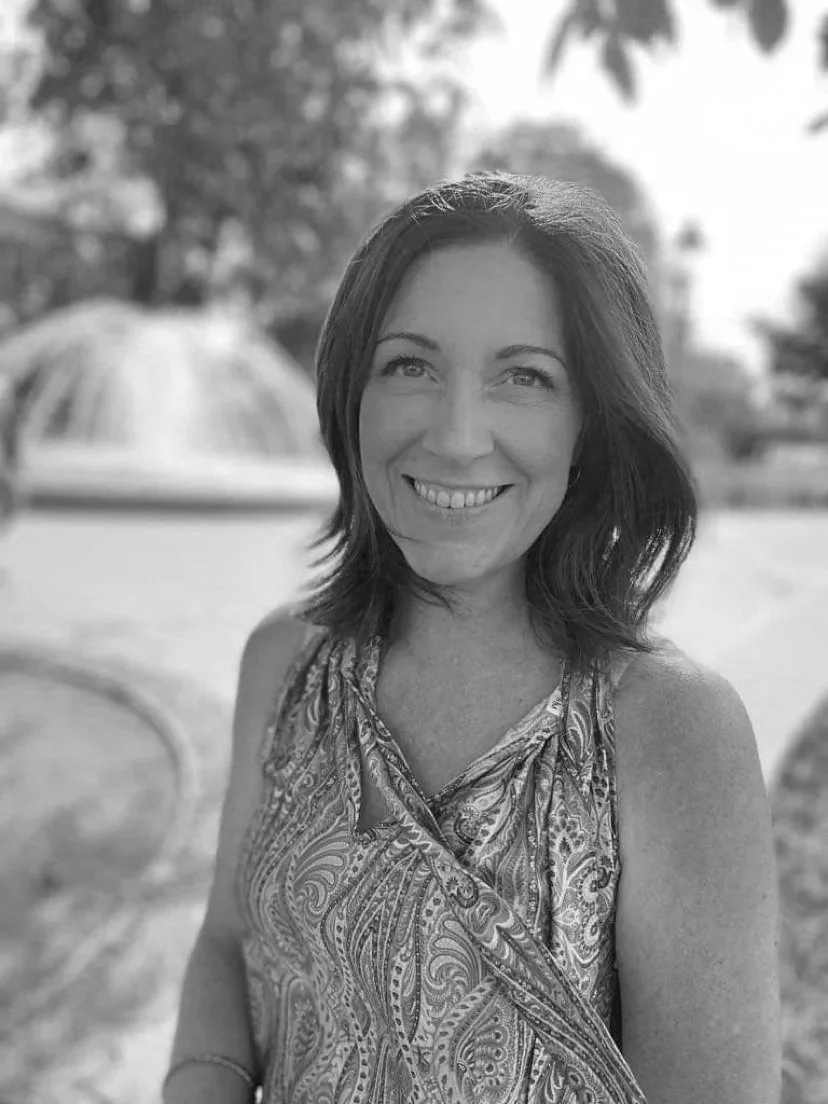 A woman with shoulder-length dark hair smiling outdoors, wearing a patterned sleeveless top, with trees and a fountain in the background.