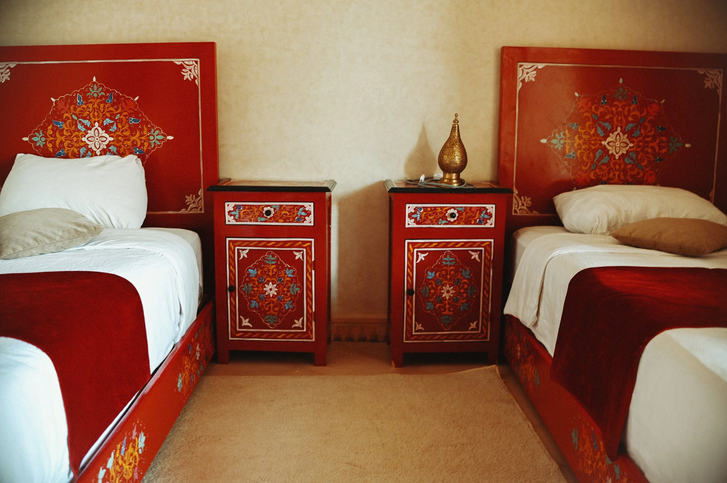 Two hotel beds with red wooden headboards and matching nightstands, decorated with intricate floral and bird designs, and a brass table lamp on the right nightstand.