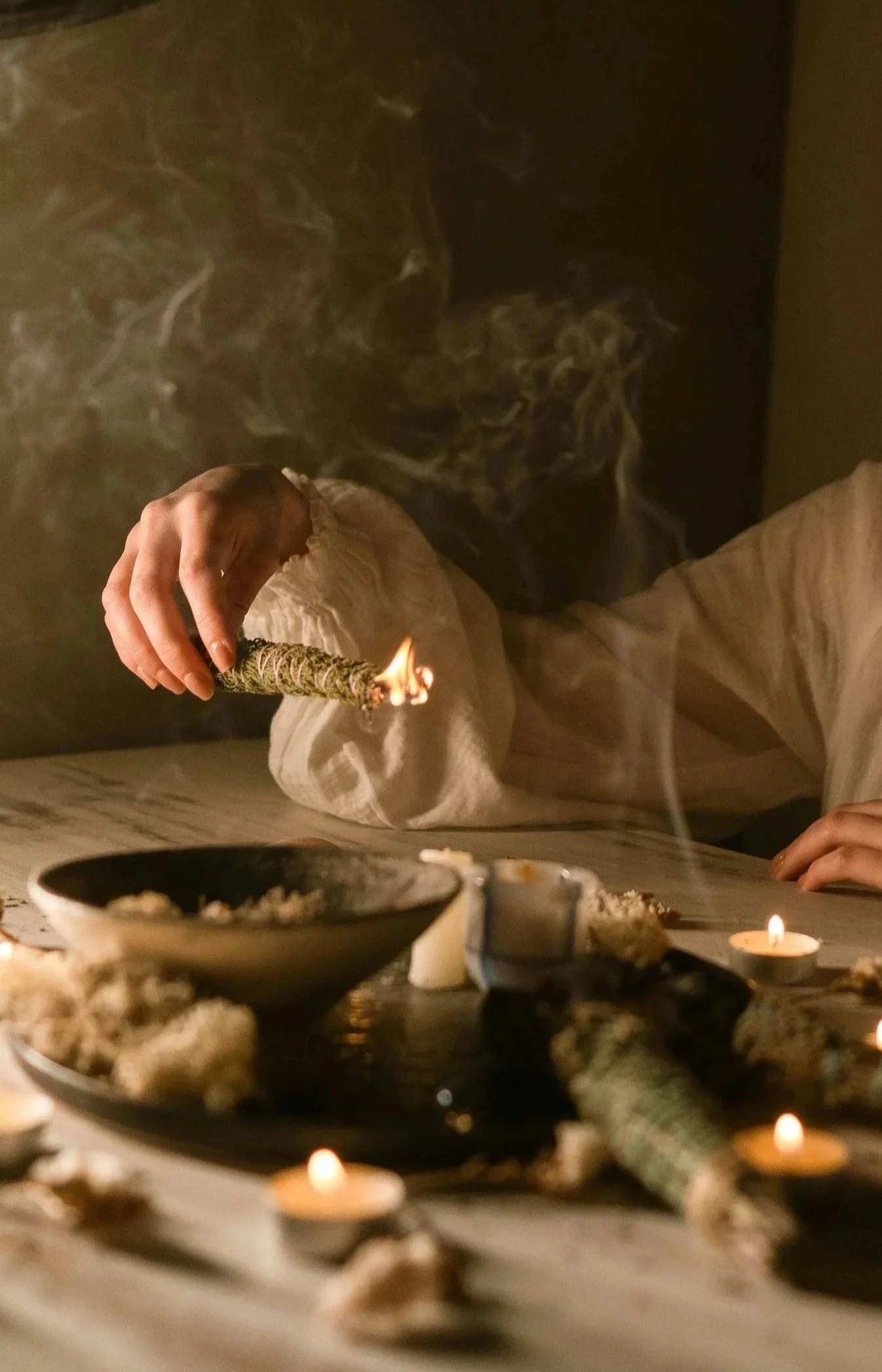 Person burning a large rolled joint in a dimly lit room with candles and bowls of herbs and incense on the table.