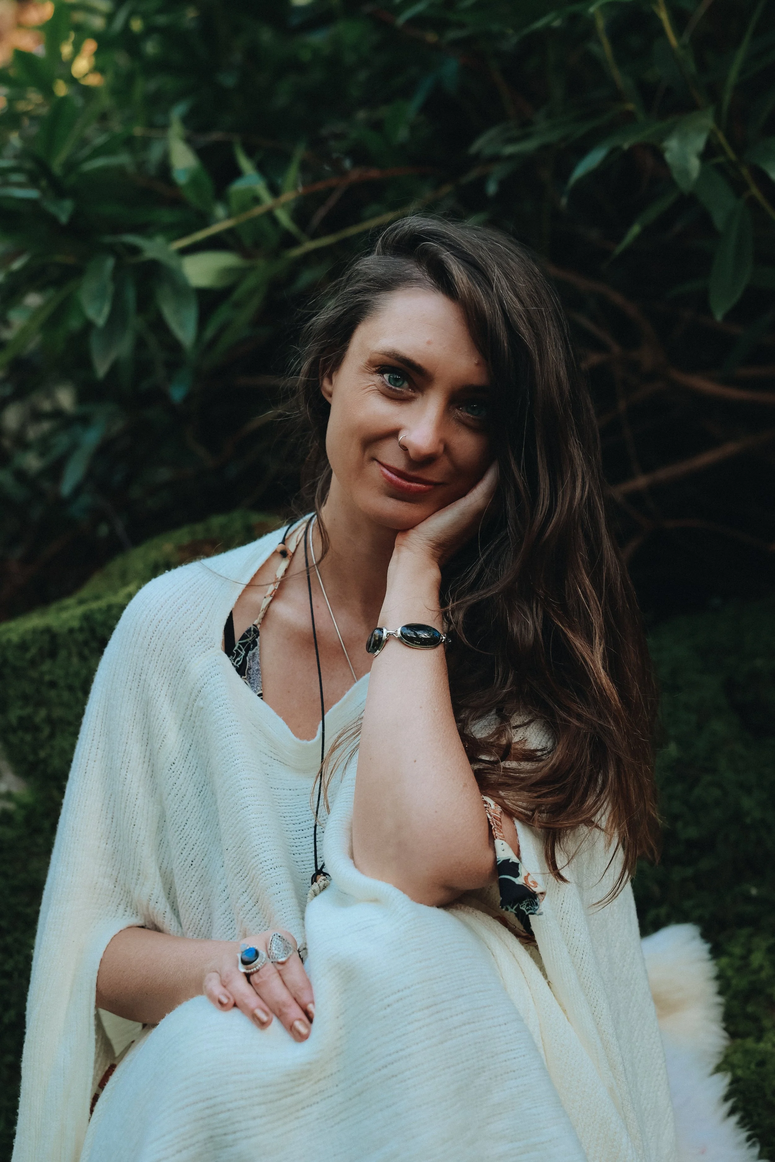 A woman with long wavy brown hair and blue eyes sitting outdoors among green bushes. She is smiling softly, resting her face on her right hand and wearing jewelry including rings, bracelets, and necklaces. She is dressed in a cream-colored cardigan over a patterned top.