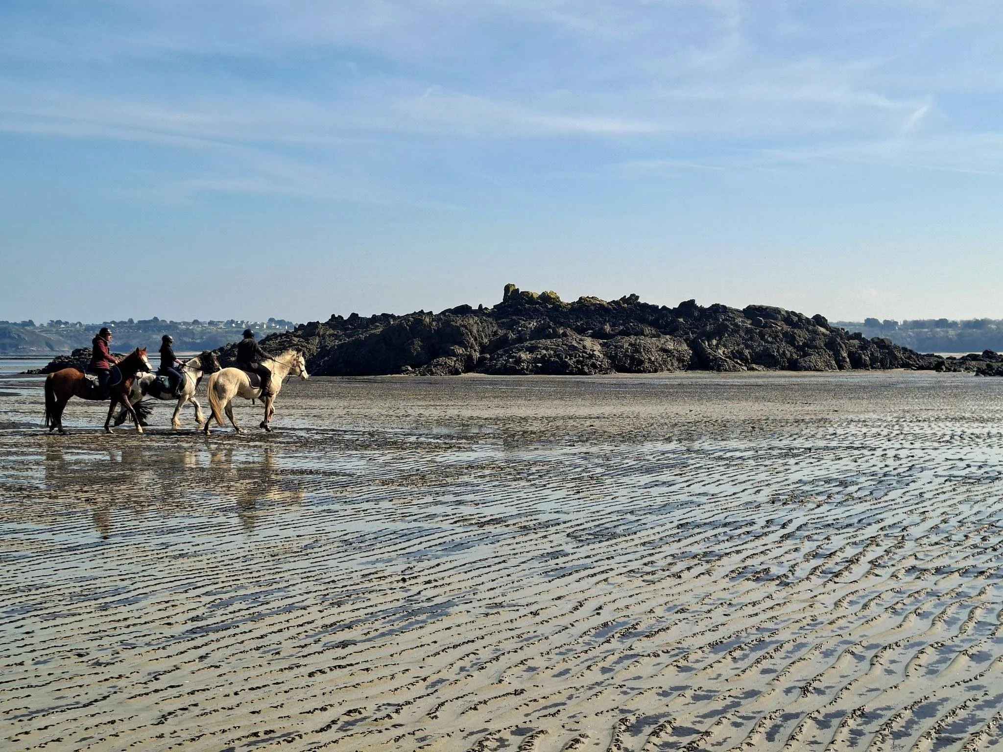 Quatre personnes à cheval traversent une plage de sable avec des rochers en arrière-plan, sous un ciel bleu clair avec quelques nuages.