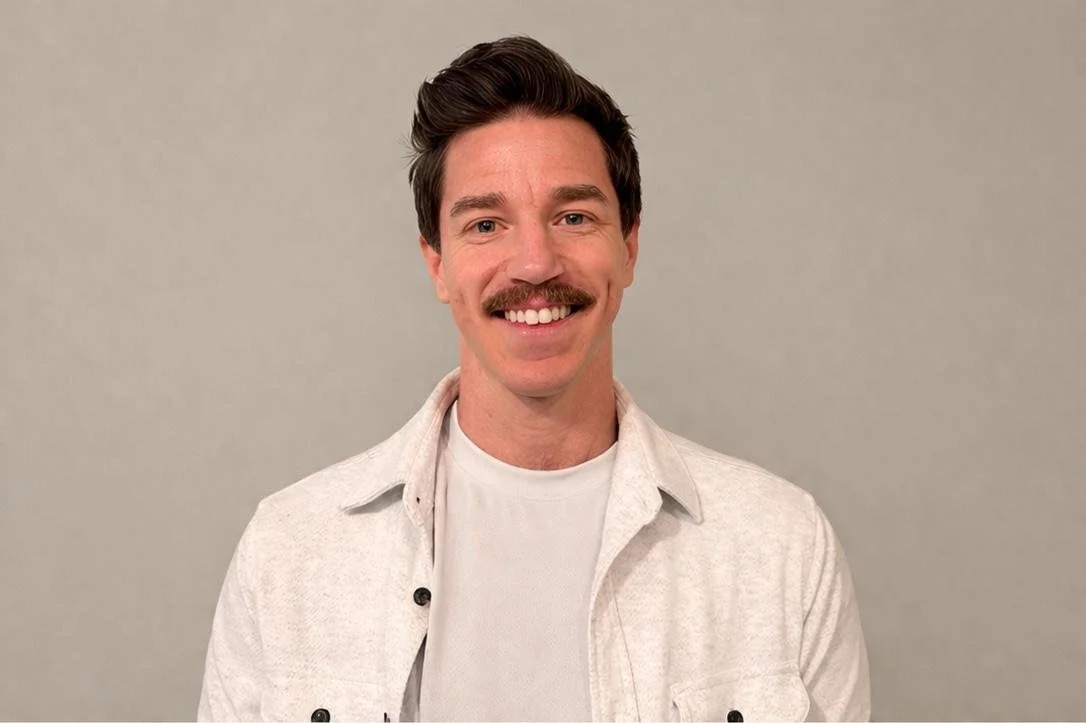 Smiling man with brown hair, mustache, wearing a white shirt and light jacket, standing against a plain gray background.