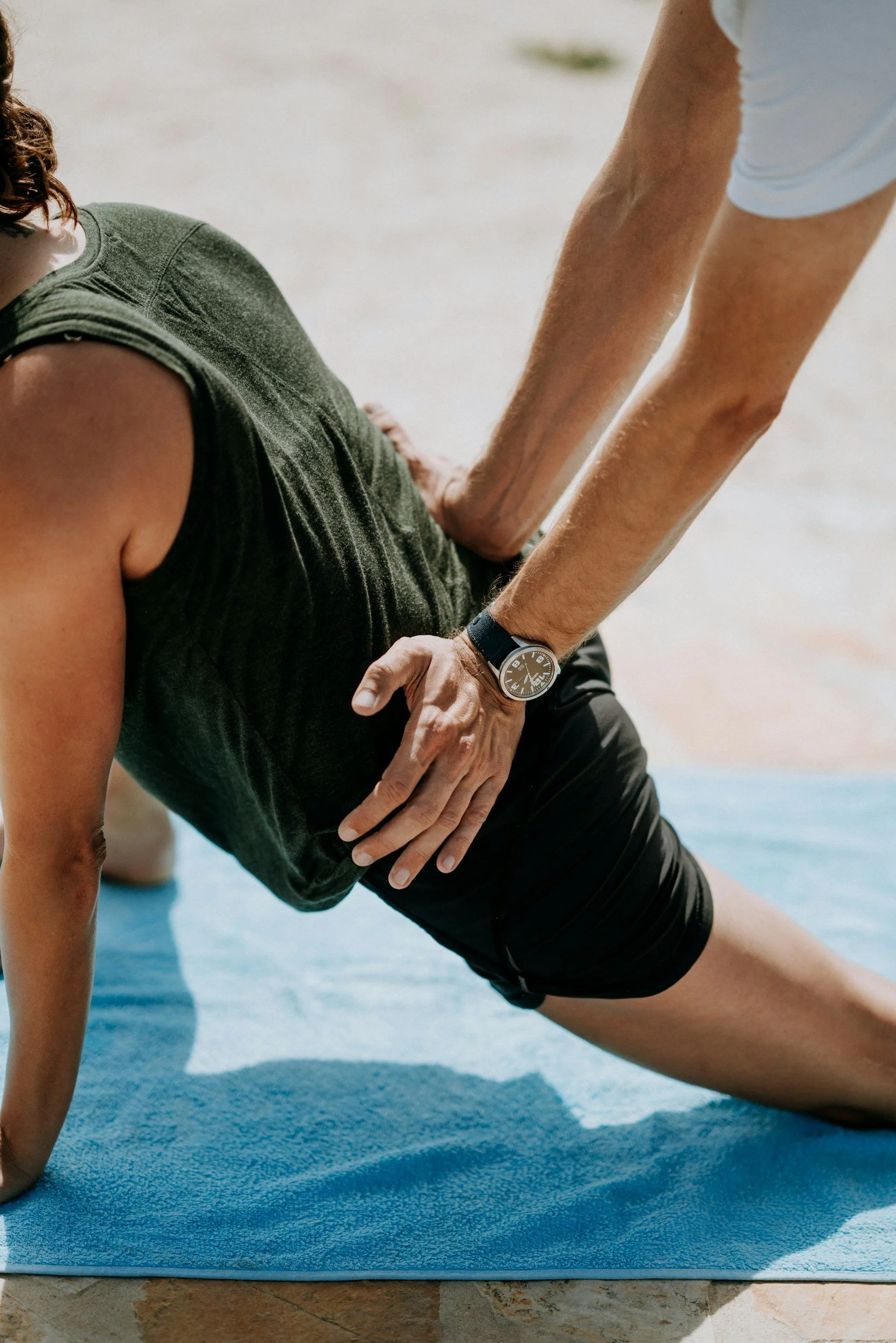A person doing a yoga pose on a blue mat, with another person supporting them by holding their side, outdoors on a sunny day.