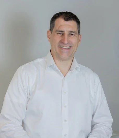 A smiling man with short dark hair wearing a white button-up shirt against a plain light grey background.