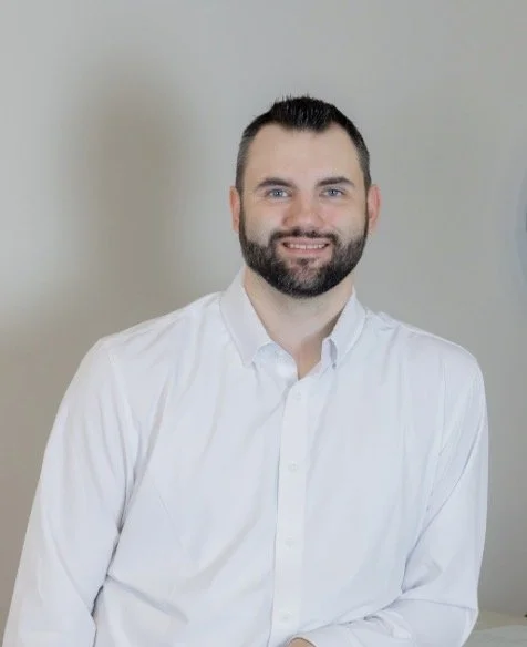 A man with a beard and short dark hair smiling at the camera, wearing a white dress shirt, standing against a plain light-colored background.