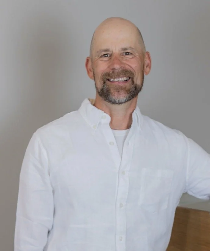 A smiling middle-aged man with a beard and bald head, wearing a white button-up shirt, standing indoors against a plain light gray backdrop.