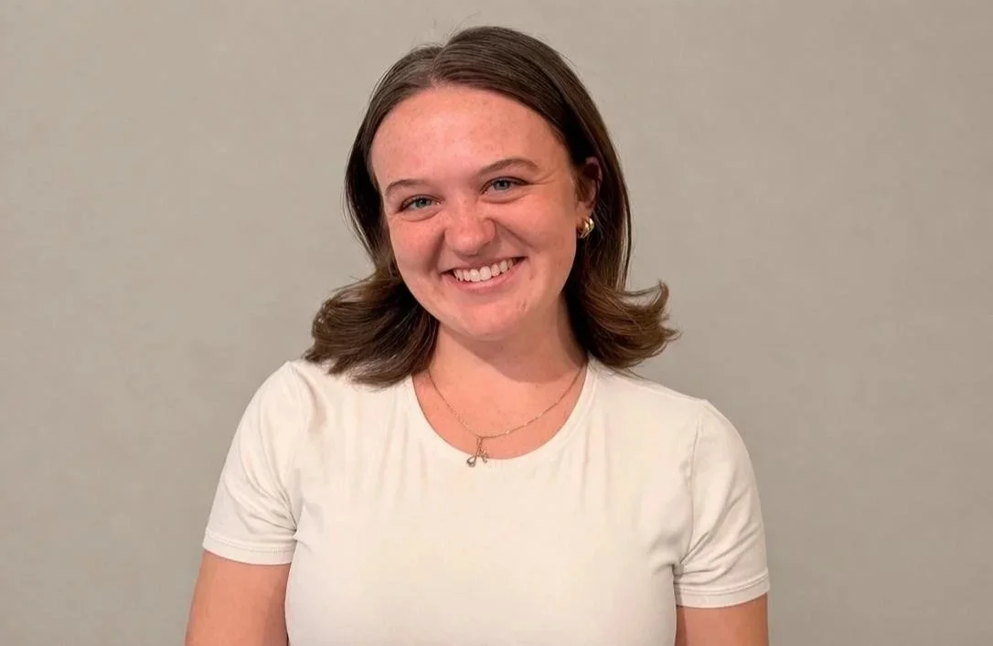 A young woman with shoulder-length brown hair smiling, wearing a white t-shirt, gold earrings, and a delicate necklace, standing against a plain gray background.