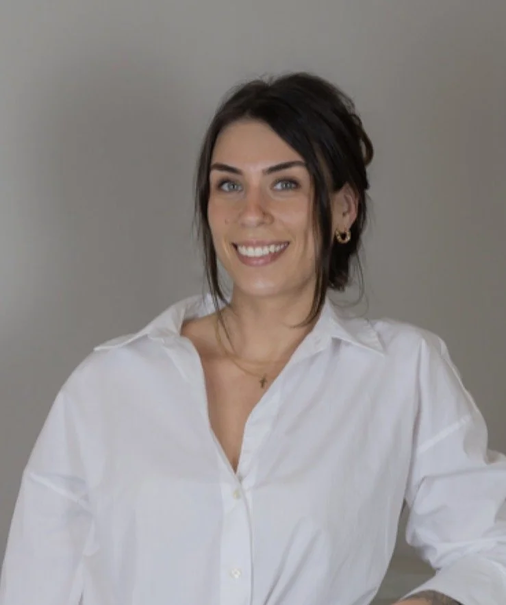 A woman with dark hair, wearing a white button-up shirt, smiling, with a neutral gray background.