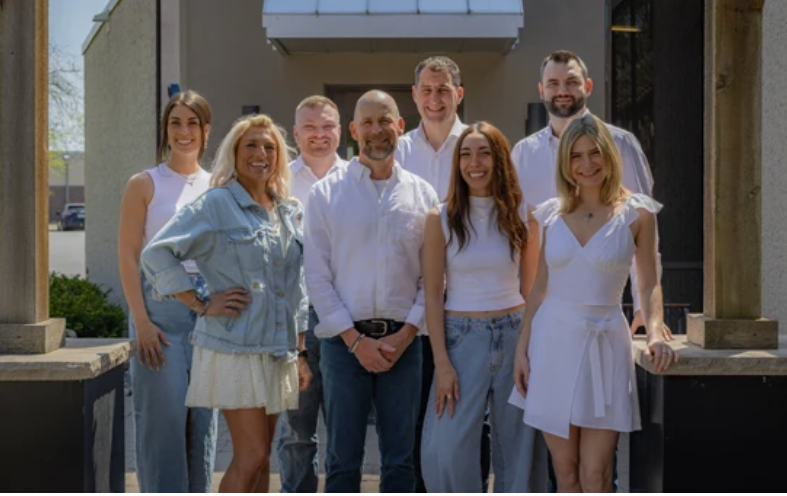 Group of seven smiling people standing outdoors in front of a building, dressed casually in white and light-colored clothing.