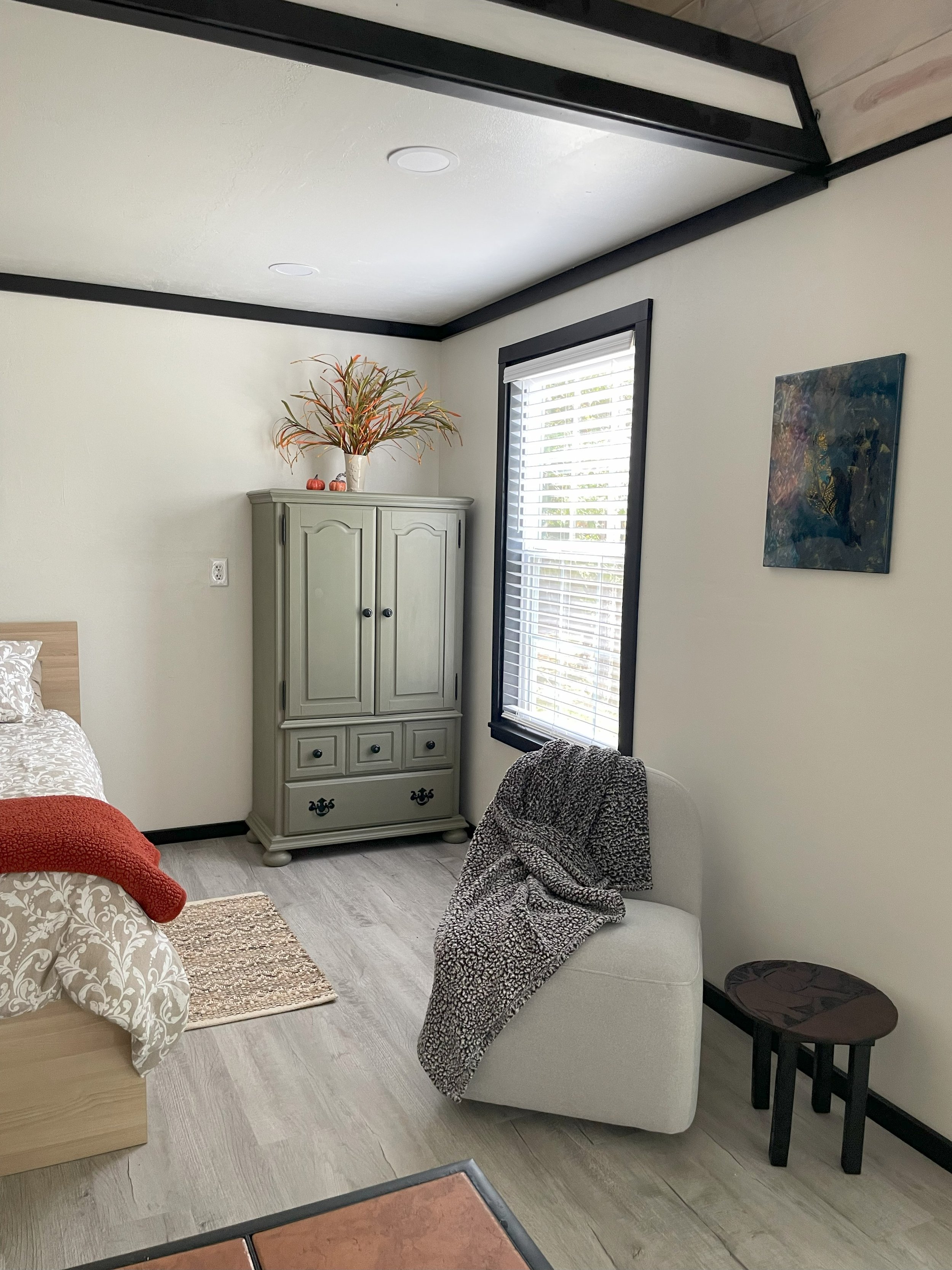 A cozy bedroom corner with a bed, a beige armchair with a patterned blanket, a small wooden side table, a green cabinet on the corner with decorative items on top, a window with blinds, a painting on the wall, and light-colored wood flooring.