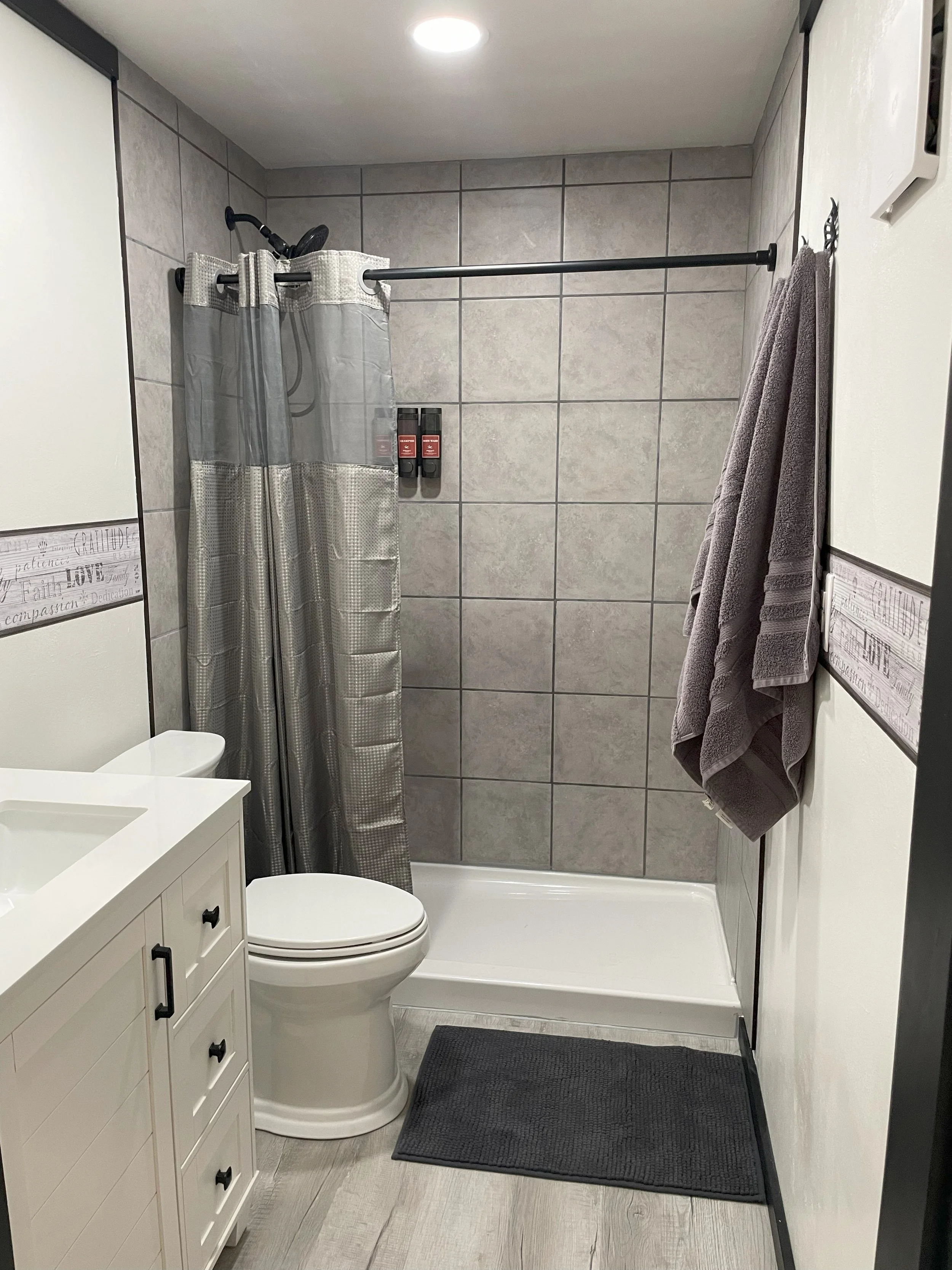 Bathroom with a shower area featuring gray tiled walls, a curtain, a black shower head, wall-mounted bottles, a white toilet, a white vanity with drawers, a gray towel hanging on a hook, a dark gray bath mat, and a ceiling light.