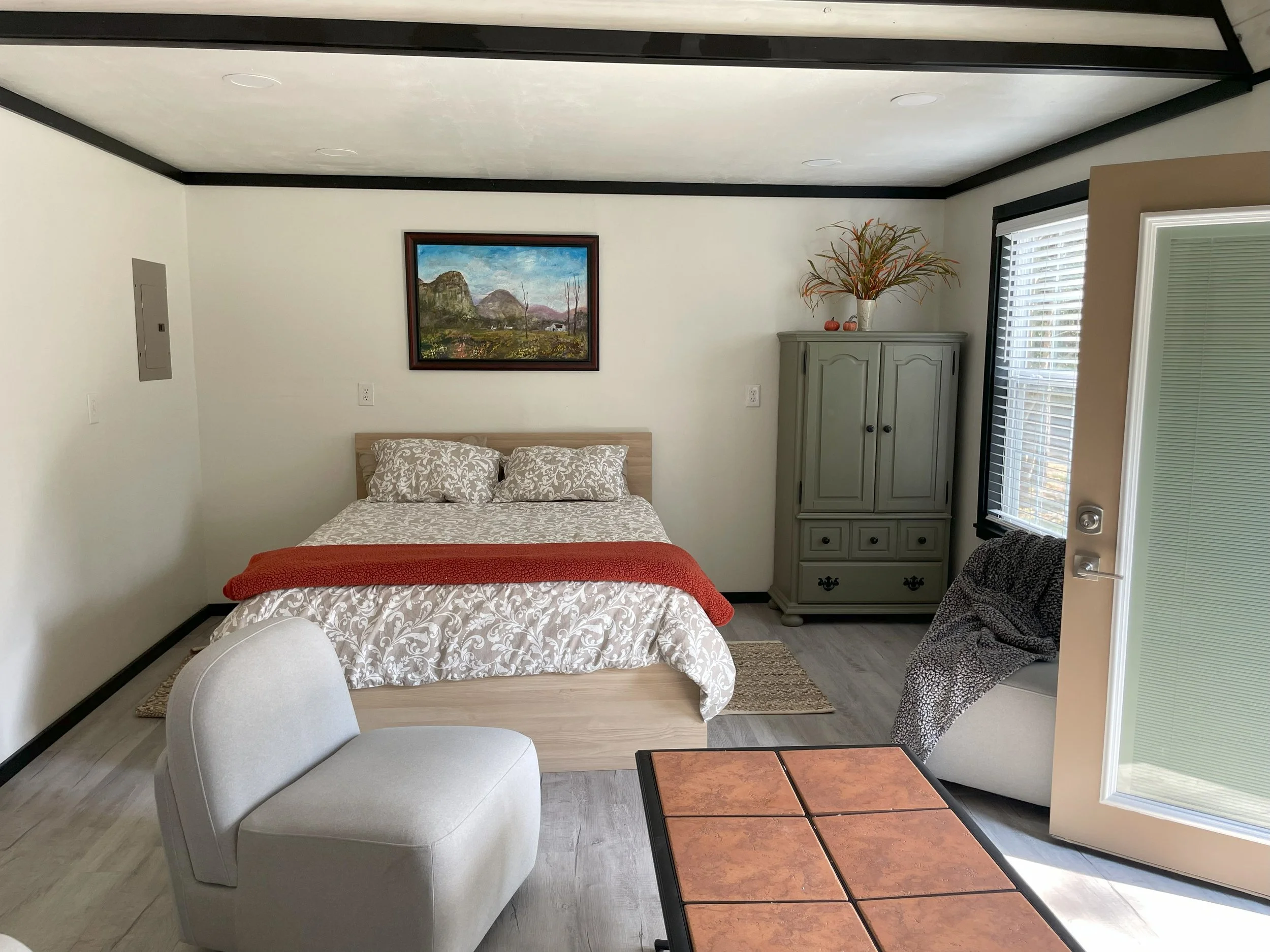 A cozy bedroom with a bed, two pillows, a framed landscape painting above, a green cabinet with decorative items, a comfortable chair with a throw blanket, a small table, and a sliding door with blinds opening to outside.