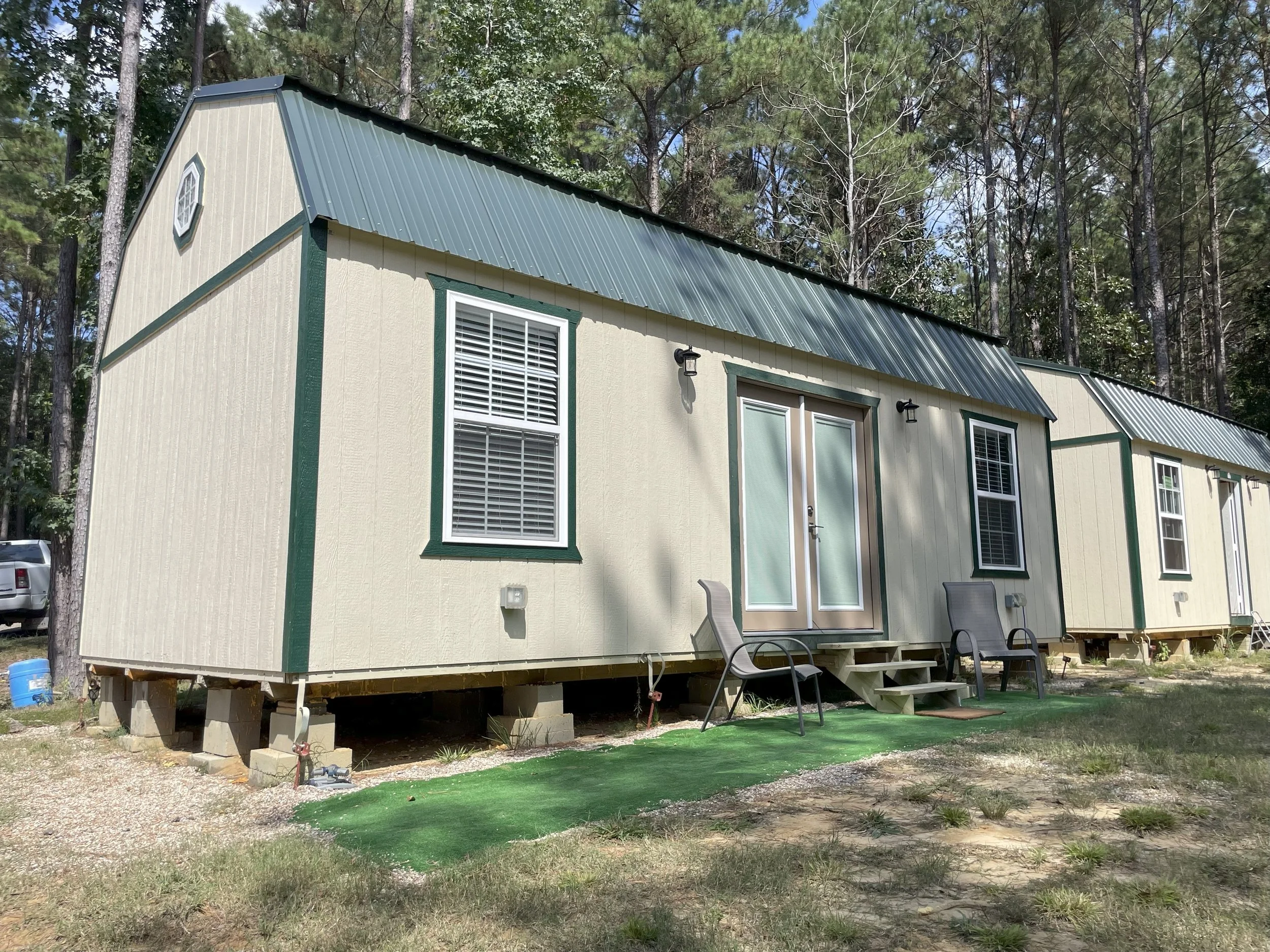Small cream-colored manufactured home with green trim, metal roof, and stairs leading to front door, surrounded by trees and outdoor furniture.