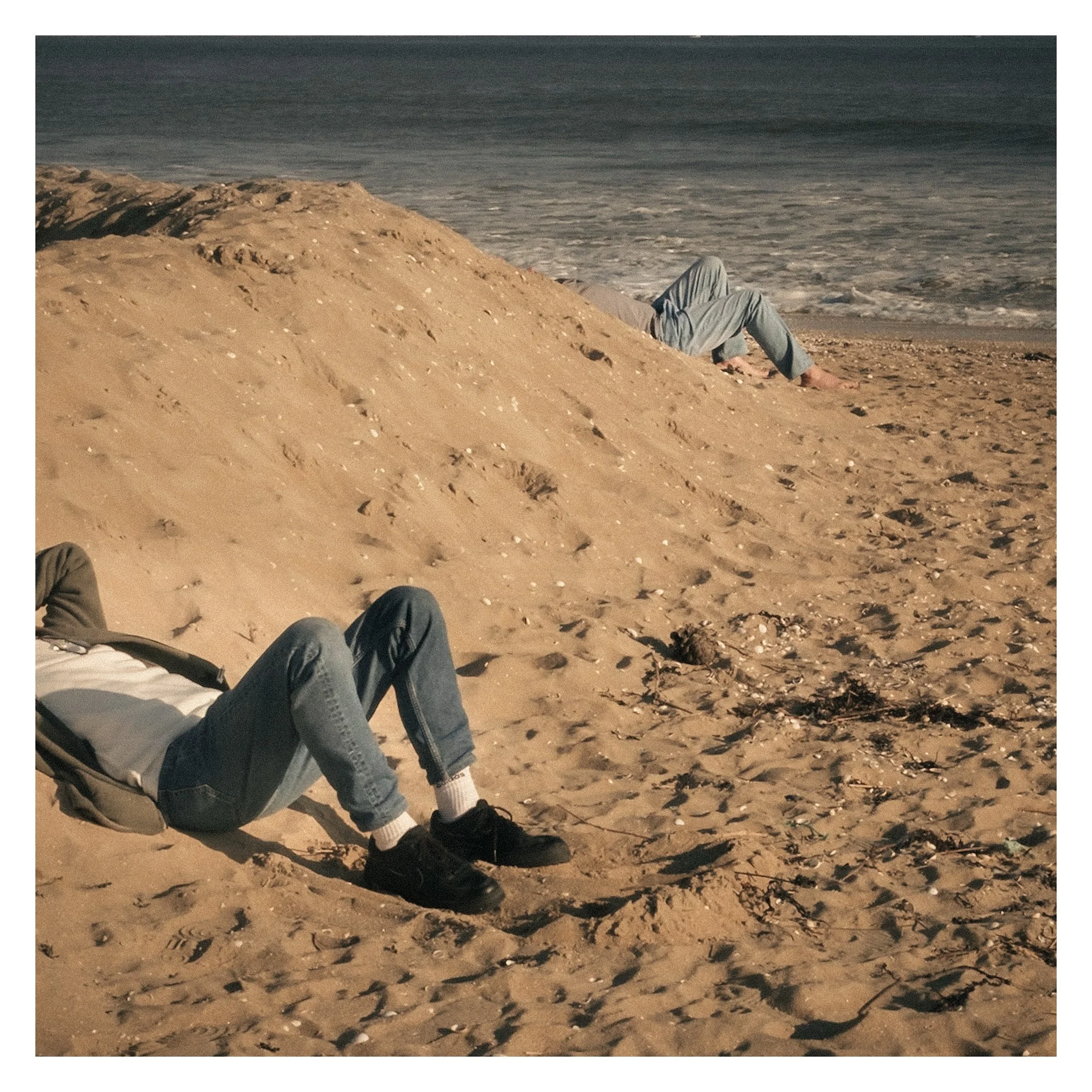 Personne allongée sur le sable d'une plage, jambes pliées, avec la mer en arrière-plan, et une autre personne allongée derrière une dune.