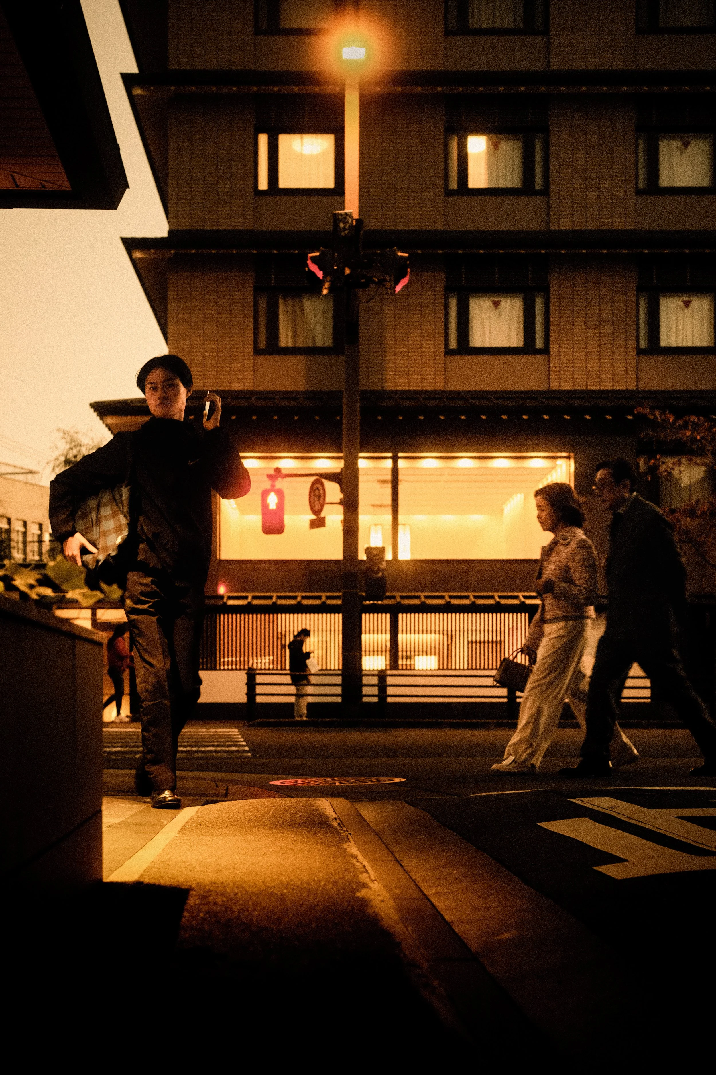 Une scène urbaine crépusculaire avec des piétons qui traversent une rue, un lampadaire illuminé au centre et un bâtiment en arrière-plan avec des fenêtres éclairées.