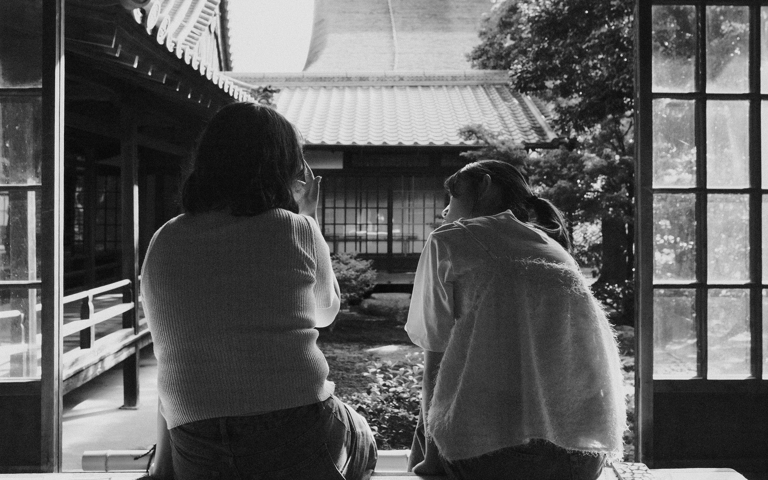 Deux femmes regardent par une fenêtre, une en utilisant des lunettes, dans un cadre traditionnel japonais en noir et blanc.