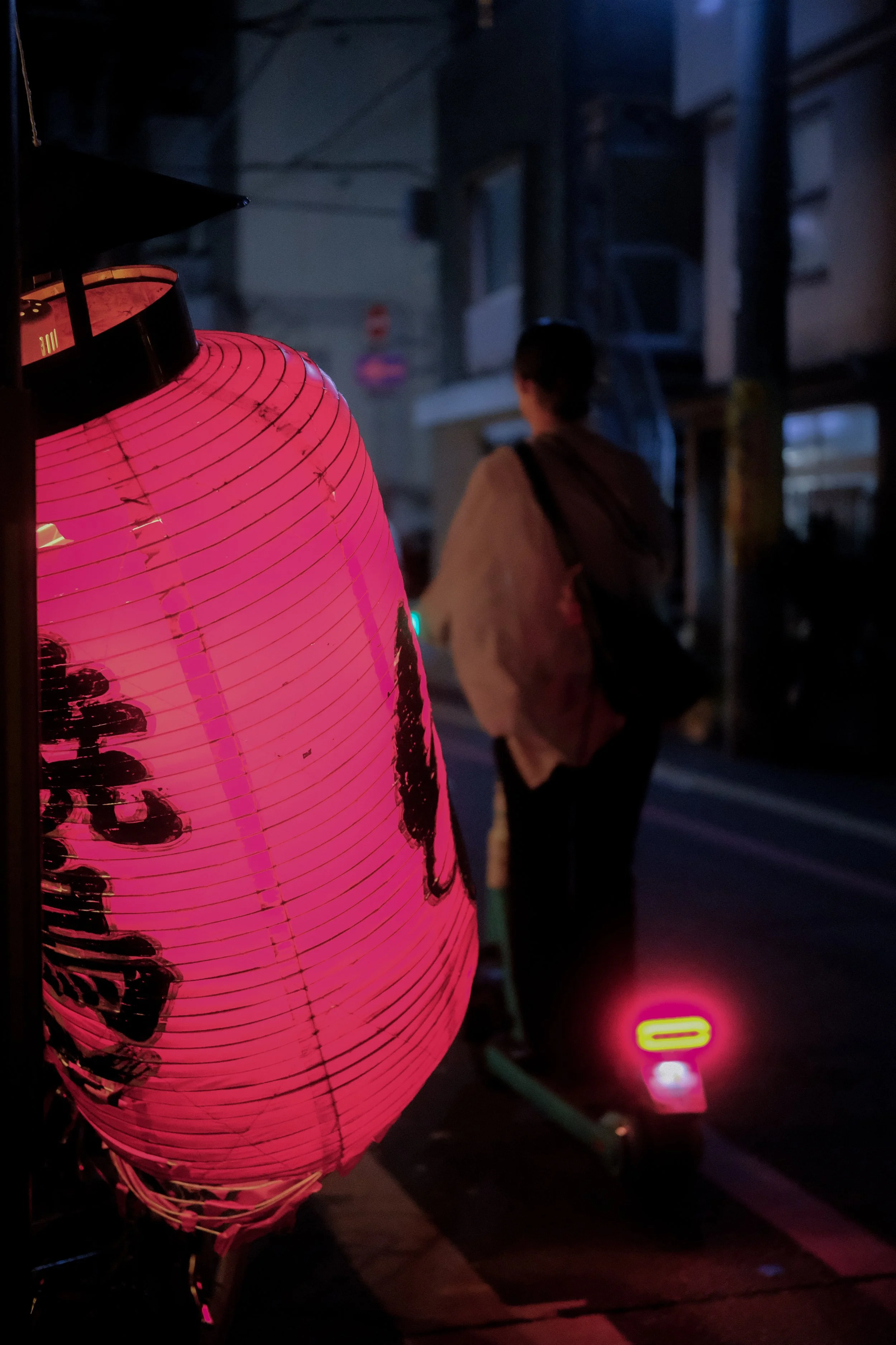 Une lanterne en papier rose illuminée, avec des caractères ou des motifs noirs, est en premier plan. En arrière-plan, une personne marchant ou attendant dans une rue nocturne, près d'une trottinette électrique émettant une lumière rose.