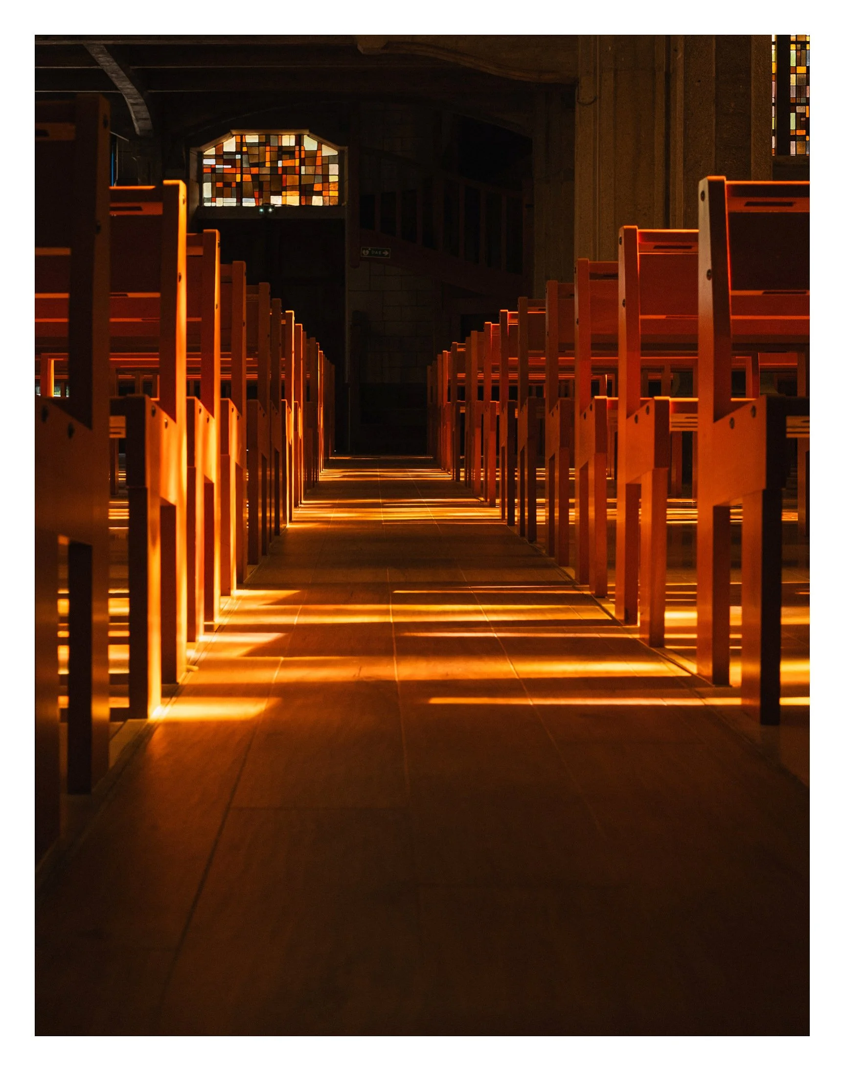 Intérieur d'une église avec des rangées de chaises en bois, éclairées par la lumière naturelle filtrant à travers des vitraux colorés.