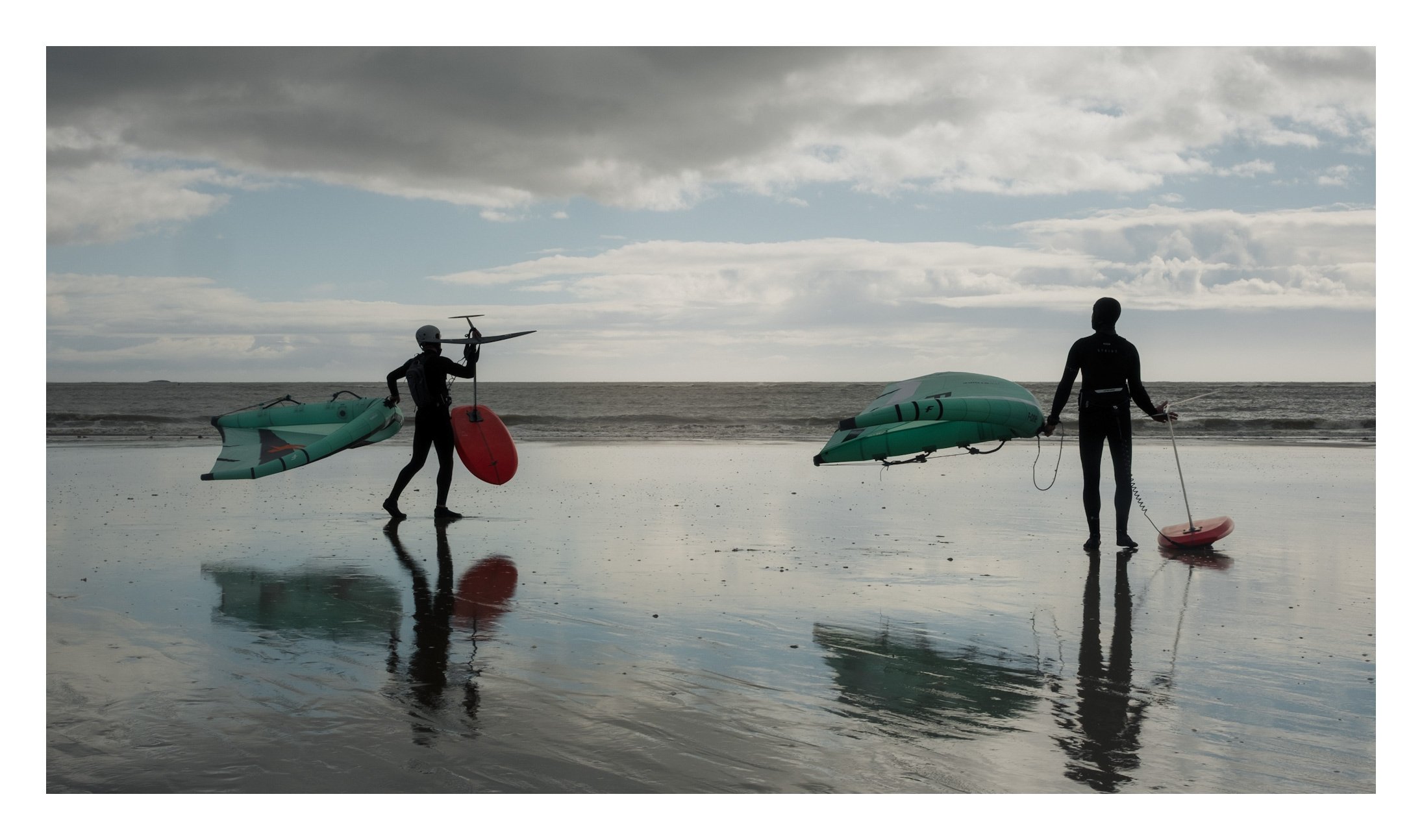 Deux personnes habillées en combinaisons de surf se tiennent sur la plage avec des planches de surf gonflables et des jet-skis, sous un ciel nuageux reflété sur le sable humide.