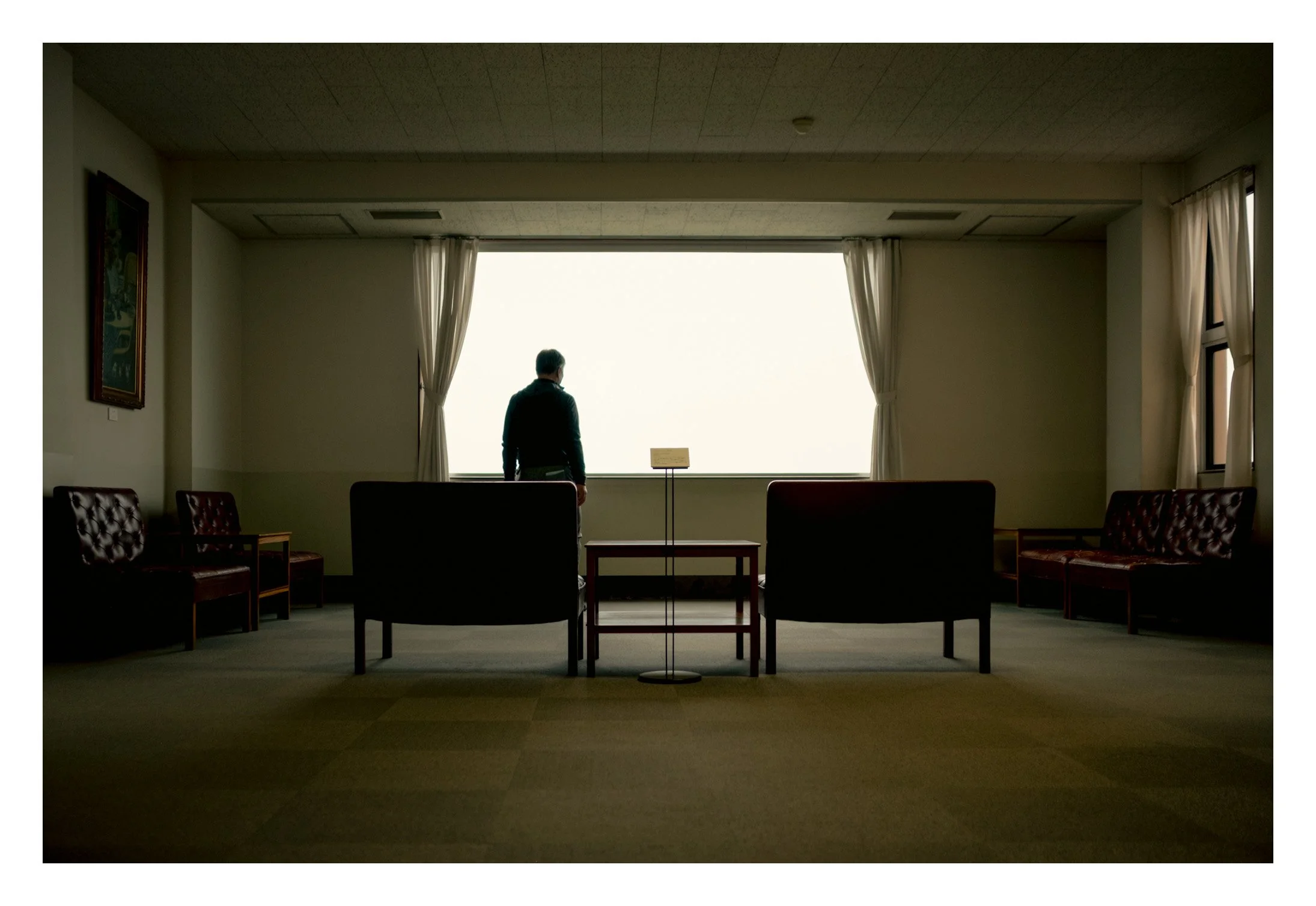 Une personne debout, observant par une grande fenêtre dans une salle de réunion vide, avec des fauteuils en velours et des rideaux blancs. L'éclairage est faible, créant une ambiance calme.