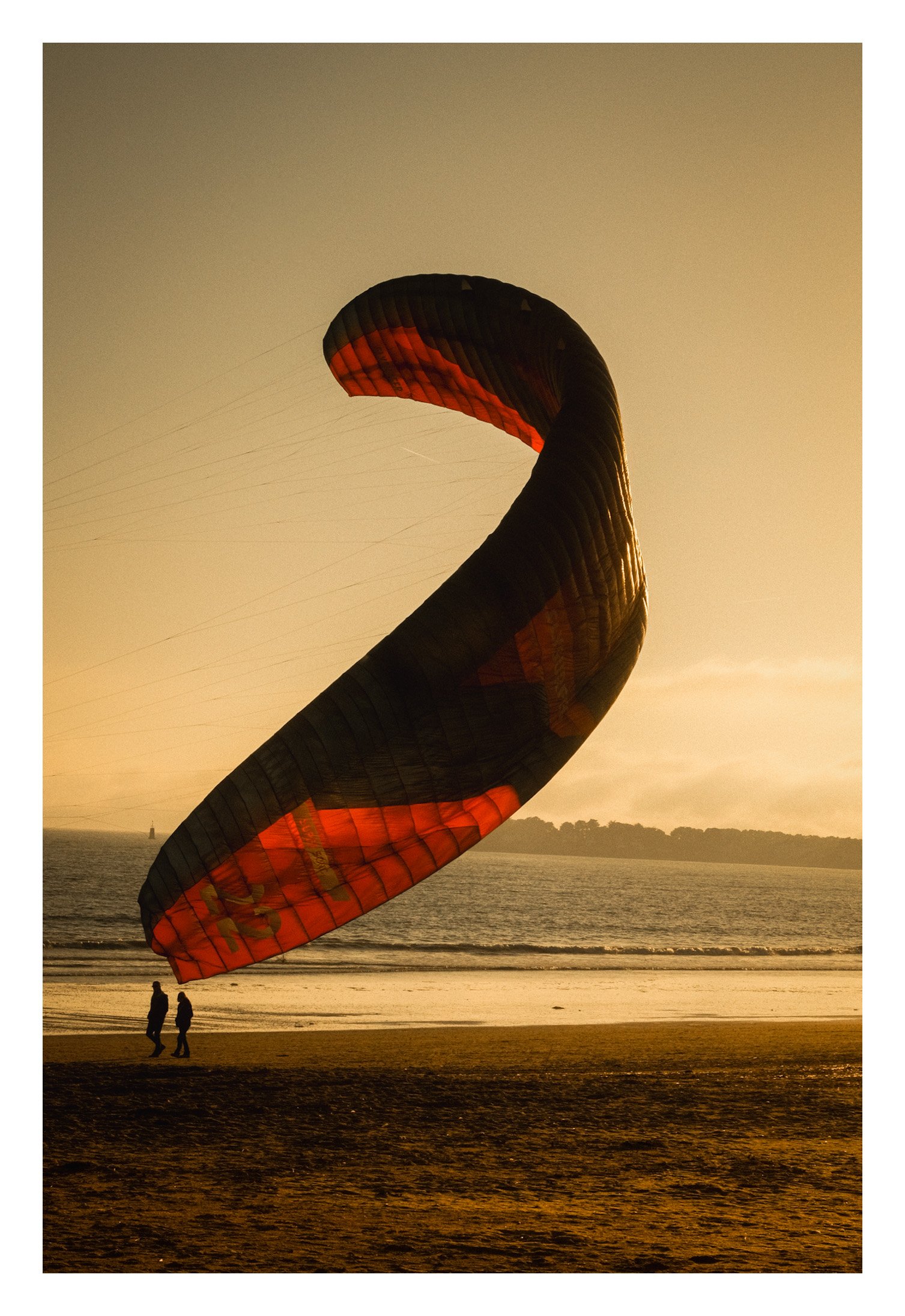 Un cerf-volant de forme incurvée, en rose et noir, vole au-dessus d'une plage au coucher du soleil, avec deux personnes qui regardent en dessous.