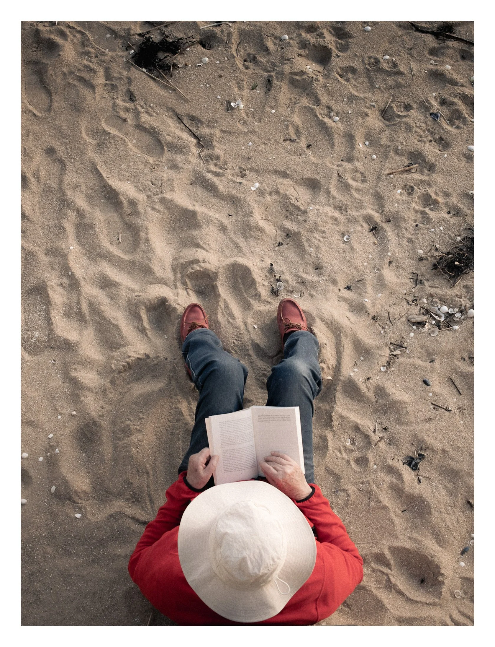 Une personne portant un chapeau blanc, un pull rouge, assise sur le sable, lit un livre avec des chaussures marron, entourée de grains de sable et de déchets Plastique.