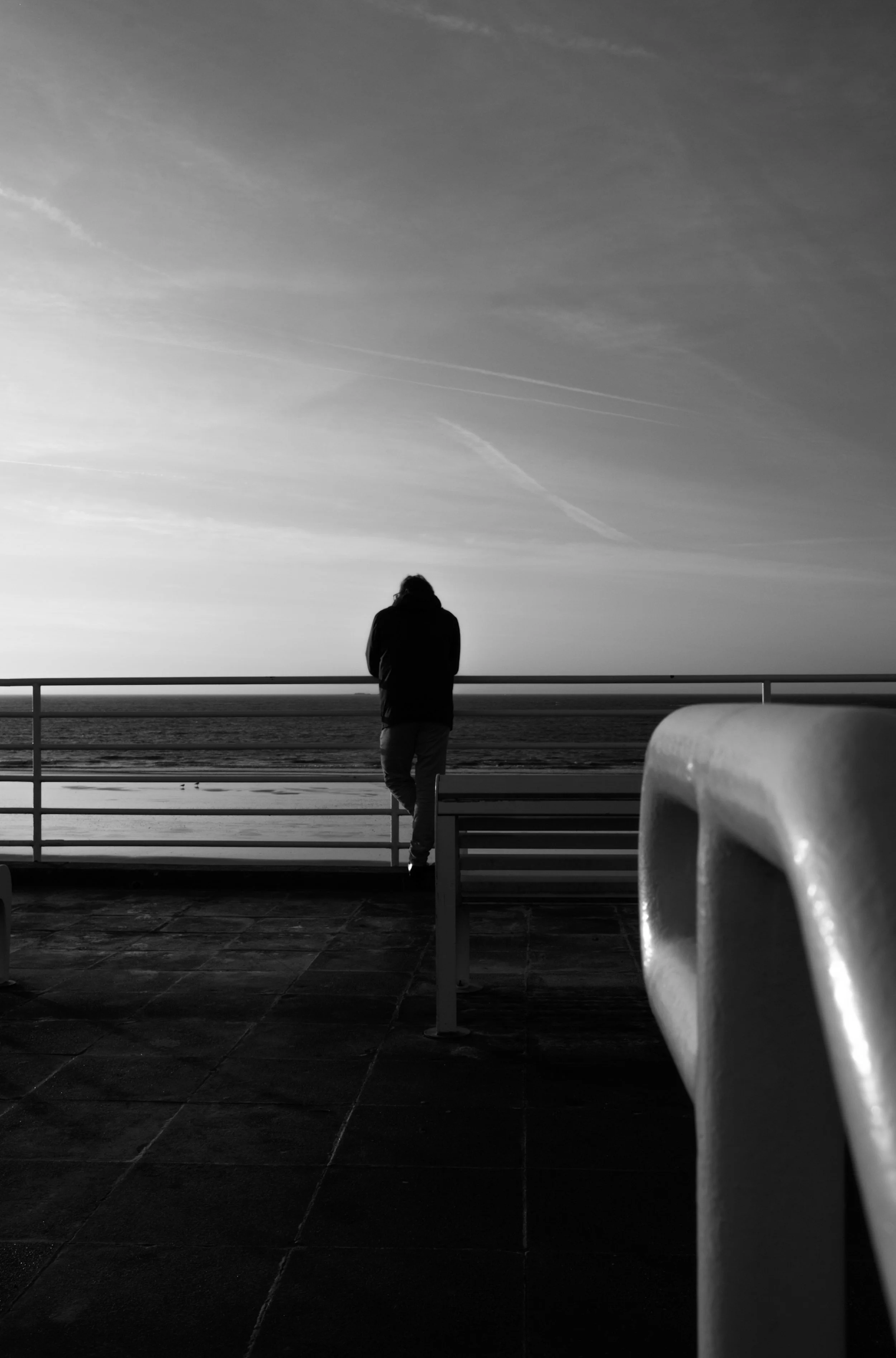 Une personne debout regardant l'océan depuis une terrasse au coucher du soleil, en noir et blanc.